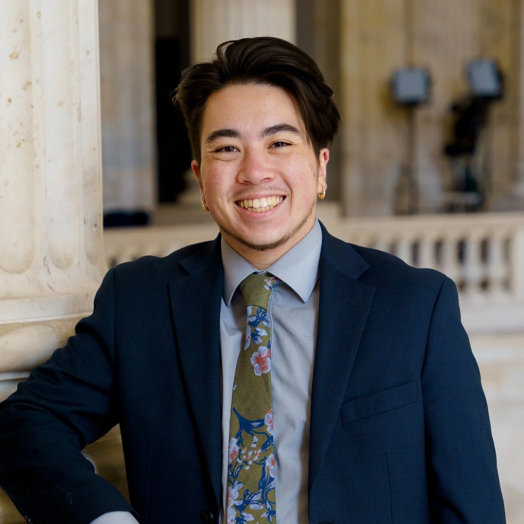 A young man with dark hair, dressed in a navy suit and floral tie, smiling confidently at the camera indoors.
