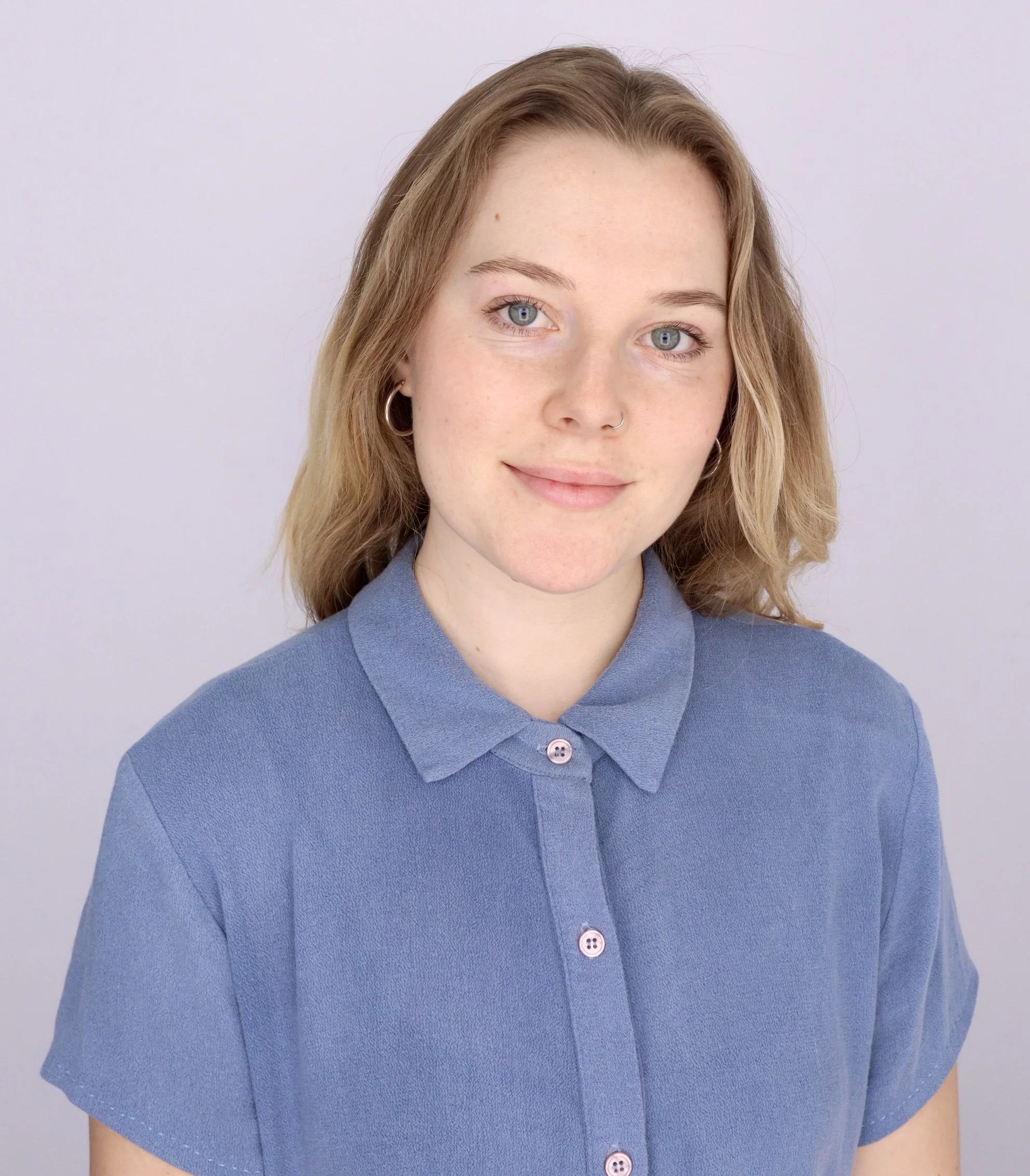 A young woman with light skin, blue eyes, and shoulder-length light brown hair, wearing a blue button-up shirt, smiling softly against a plain light background.