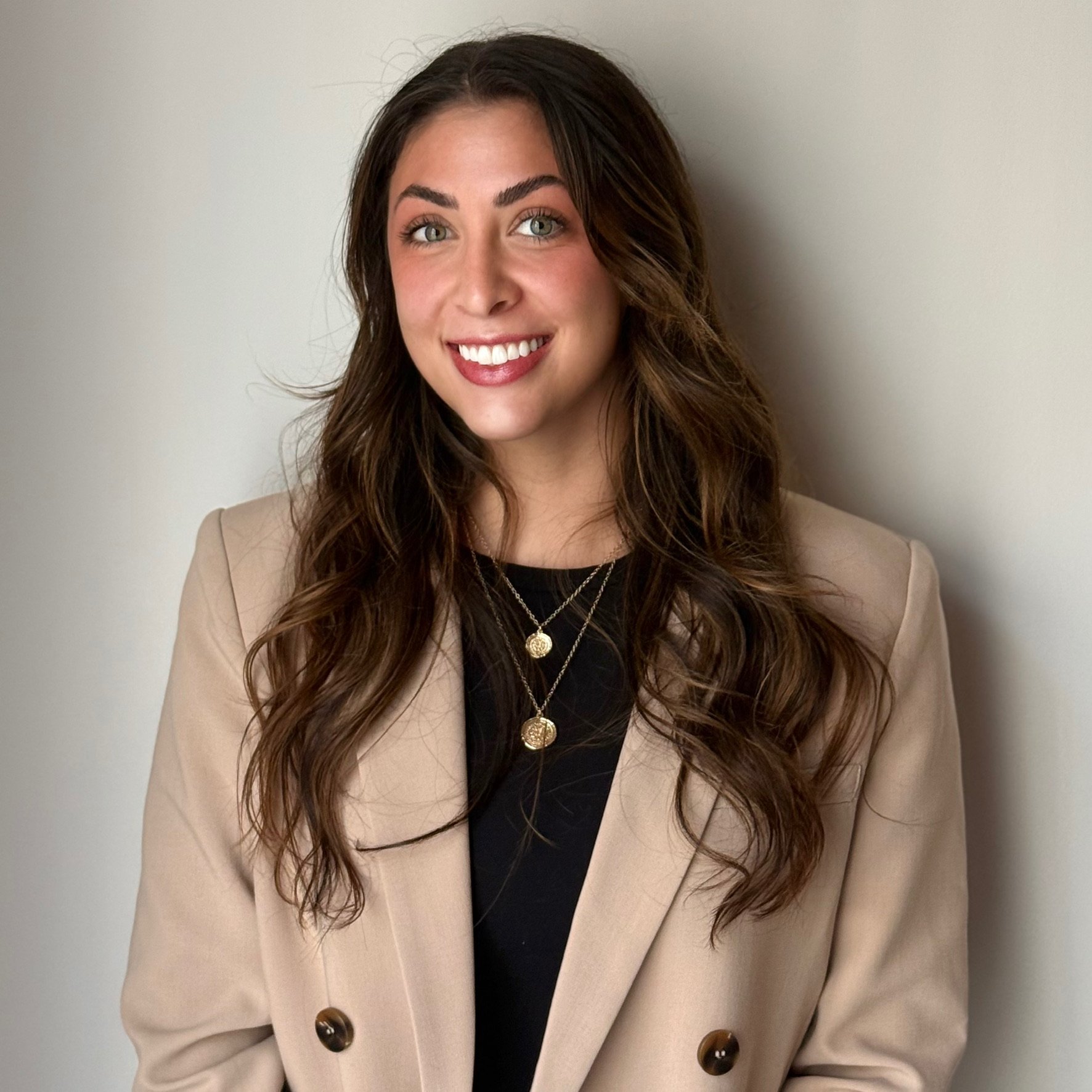A young woman with long wavy brown hair, wearing a beige blazer over a black top, smiling and standing against a plain light-colored wall.