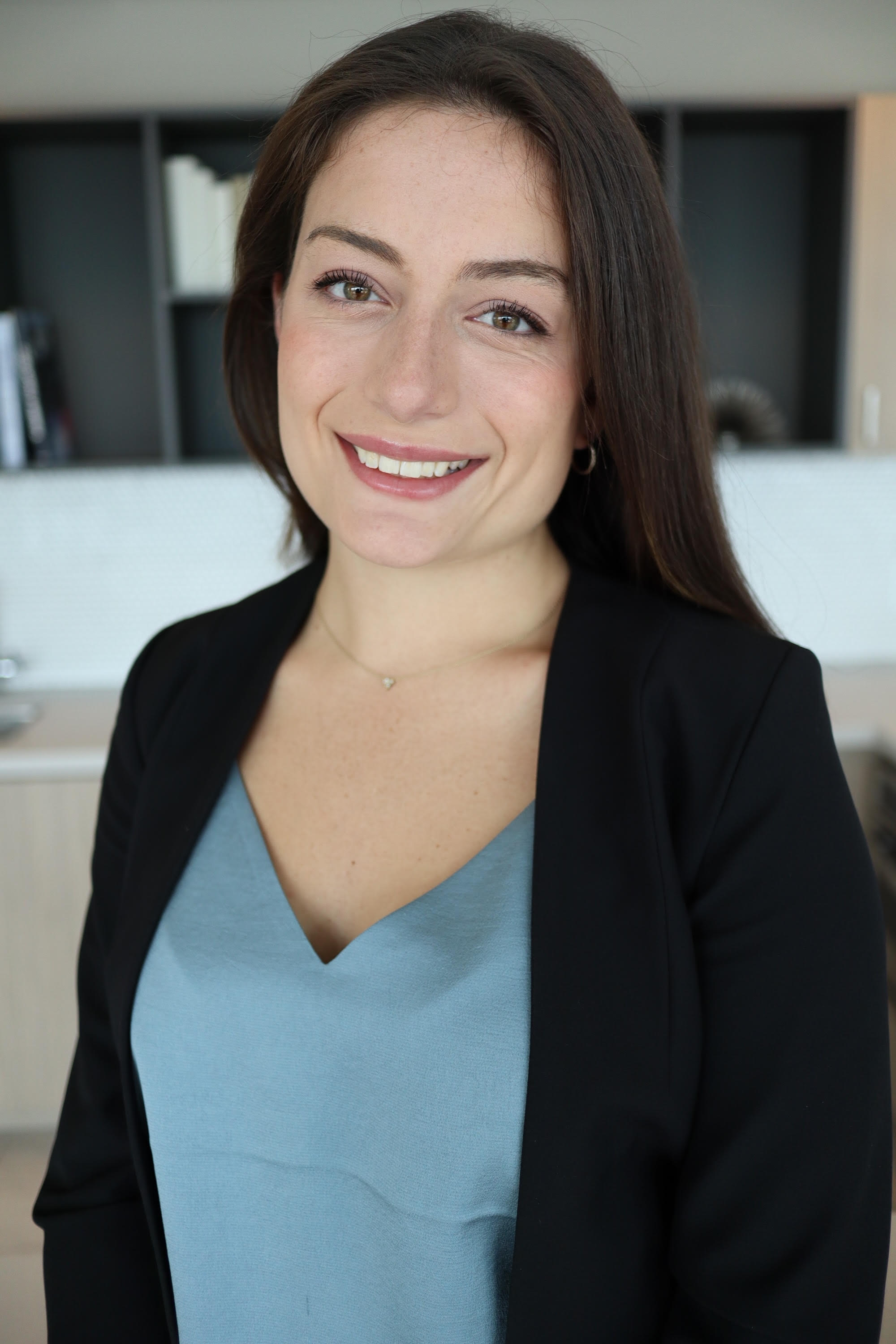 Portrait of a young woman with long brown hair, smiling, wearing a black blazer over a blue top, in an office or home setting.