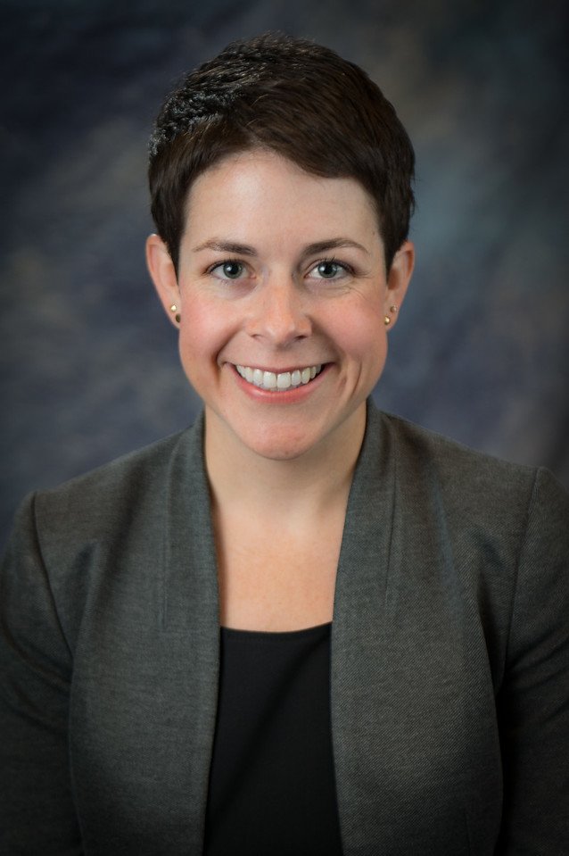 Portrait of a woman with short dark hair, wearing a dark blazer and a black top, smiling at the camera with a blurred dark background.