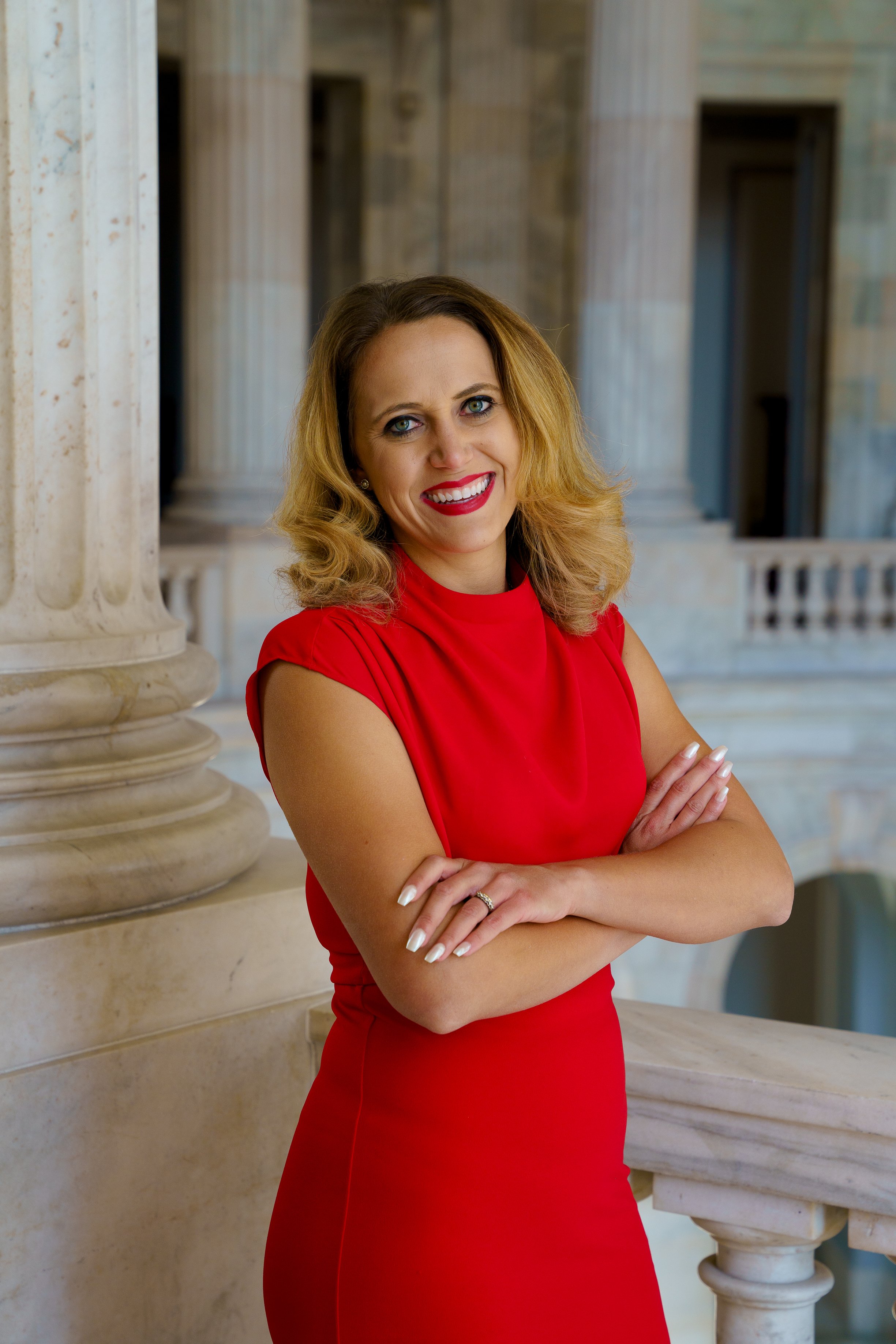 A woman with blonde hair, wearing a red dress, smiling and leaning against a marble column in a grand building with marble pillars and ornate architecture.