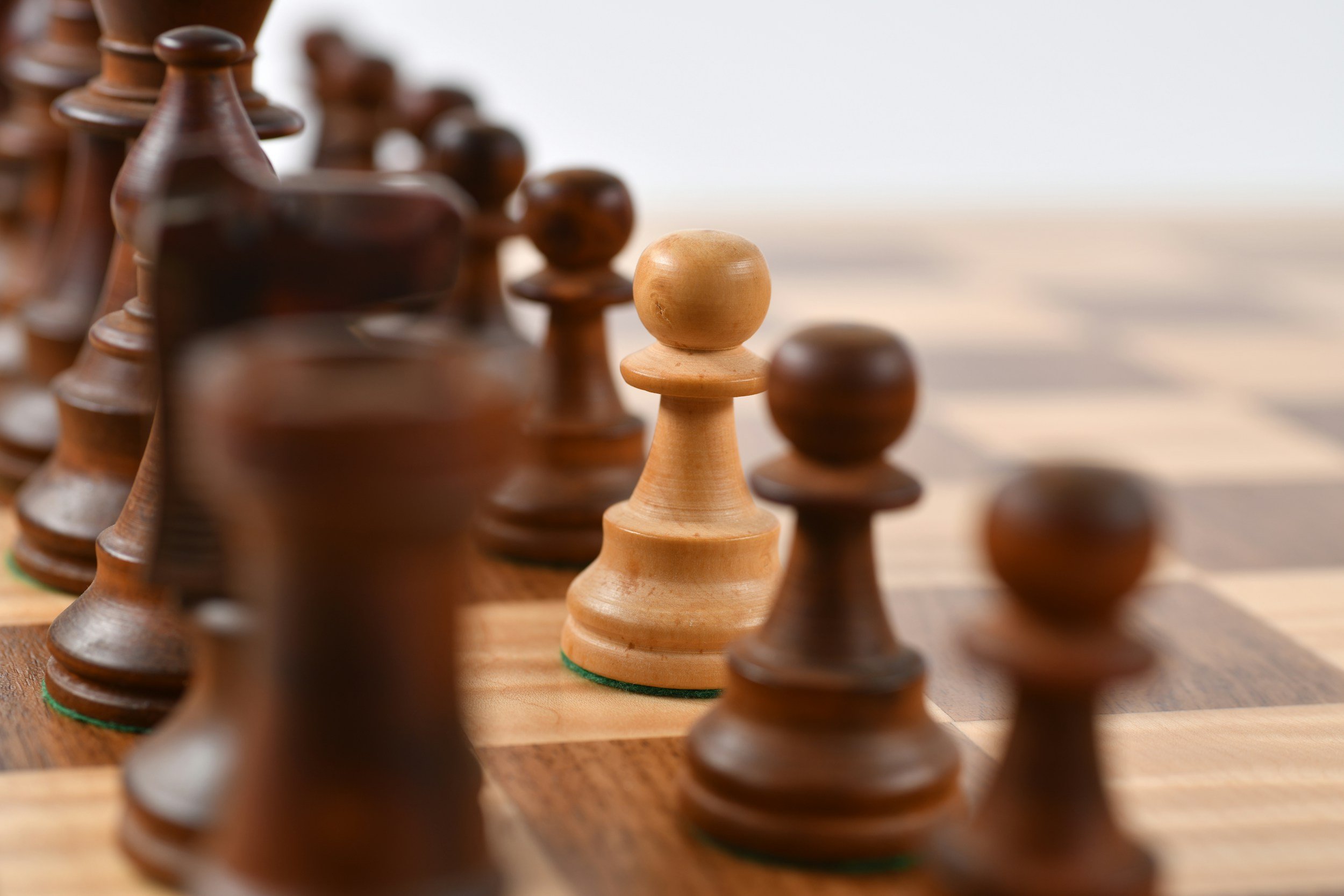 Close-up of a chessboard with wooden chess pieces, focusing on a light-colored pawn surrounded by darker pawns.