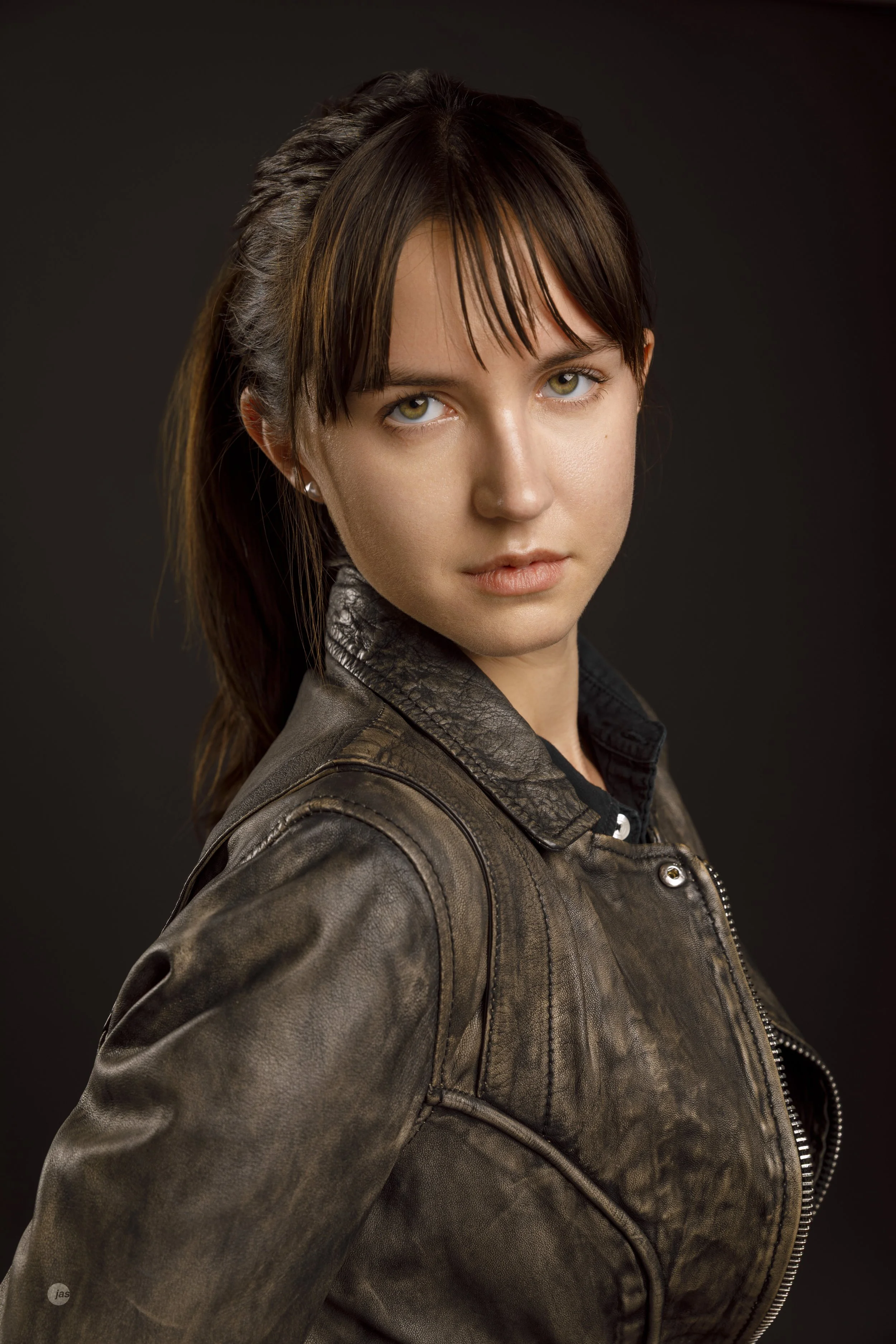 A young woman with brown hair and piercing eyes looking directly at the camera, wearing a grey leather jacket against a dark background.