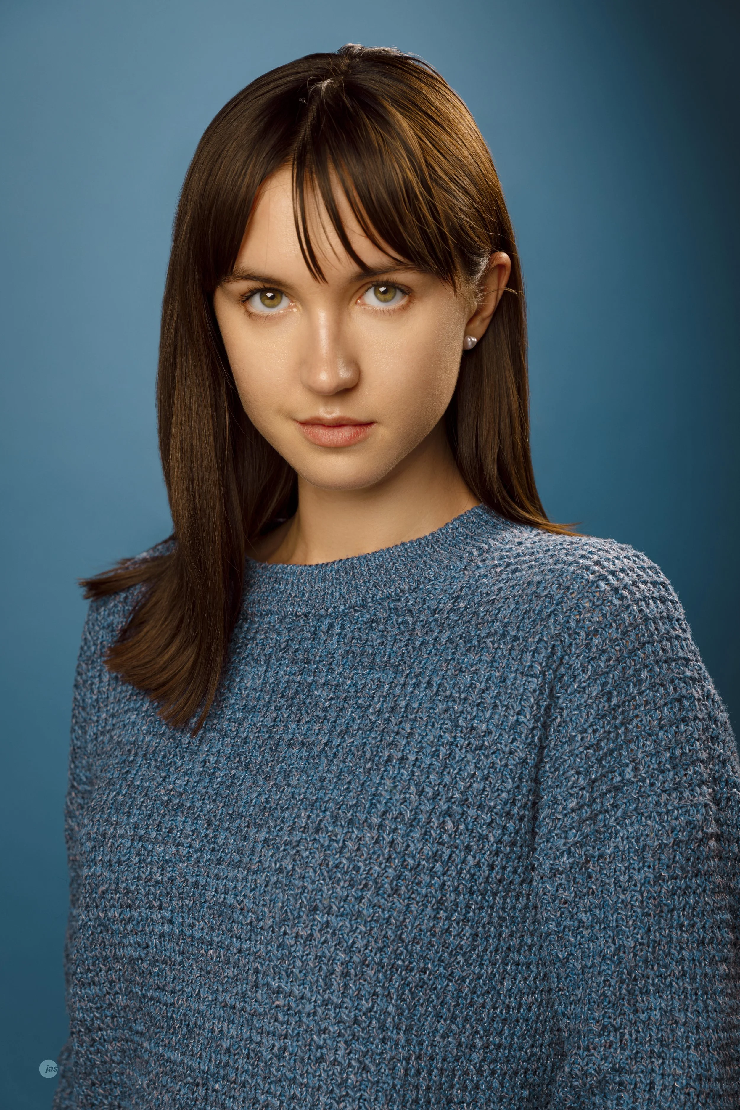 Portrait of a young woman with straight brown hair and bangs, green eyes, wearing a blue textured sweater, standing against a blue gradient background.