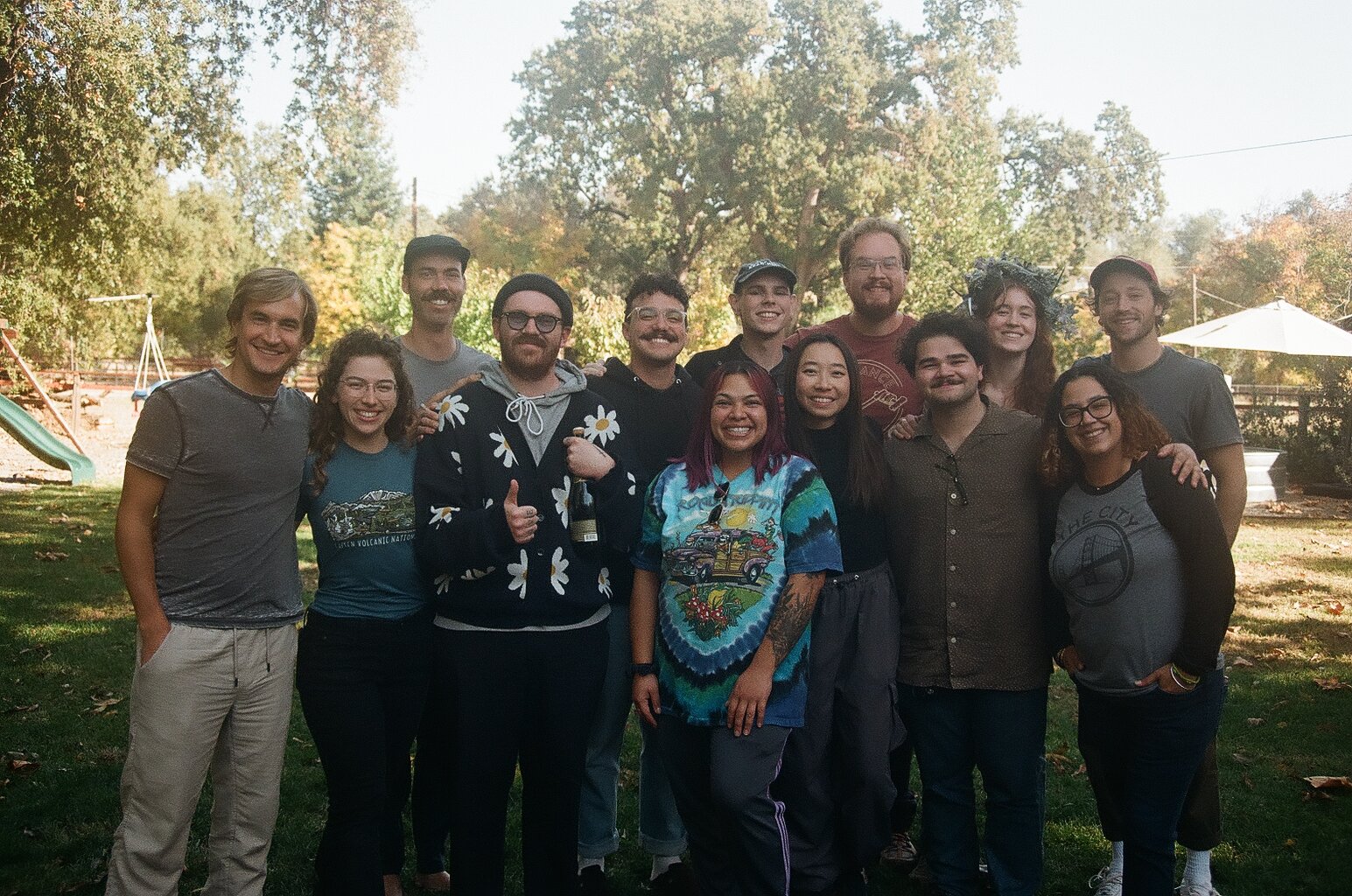 A group of cast and crew from the feature film production of The Rattlin' Bog outside in a park or backyard, smiling for a wrap party photo with trees, a slide, and an umbrella in the background.