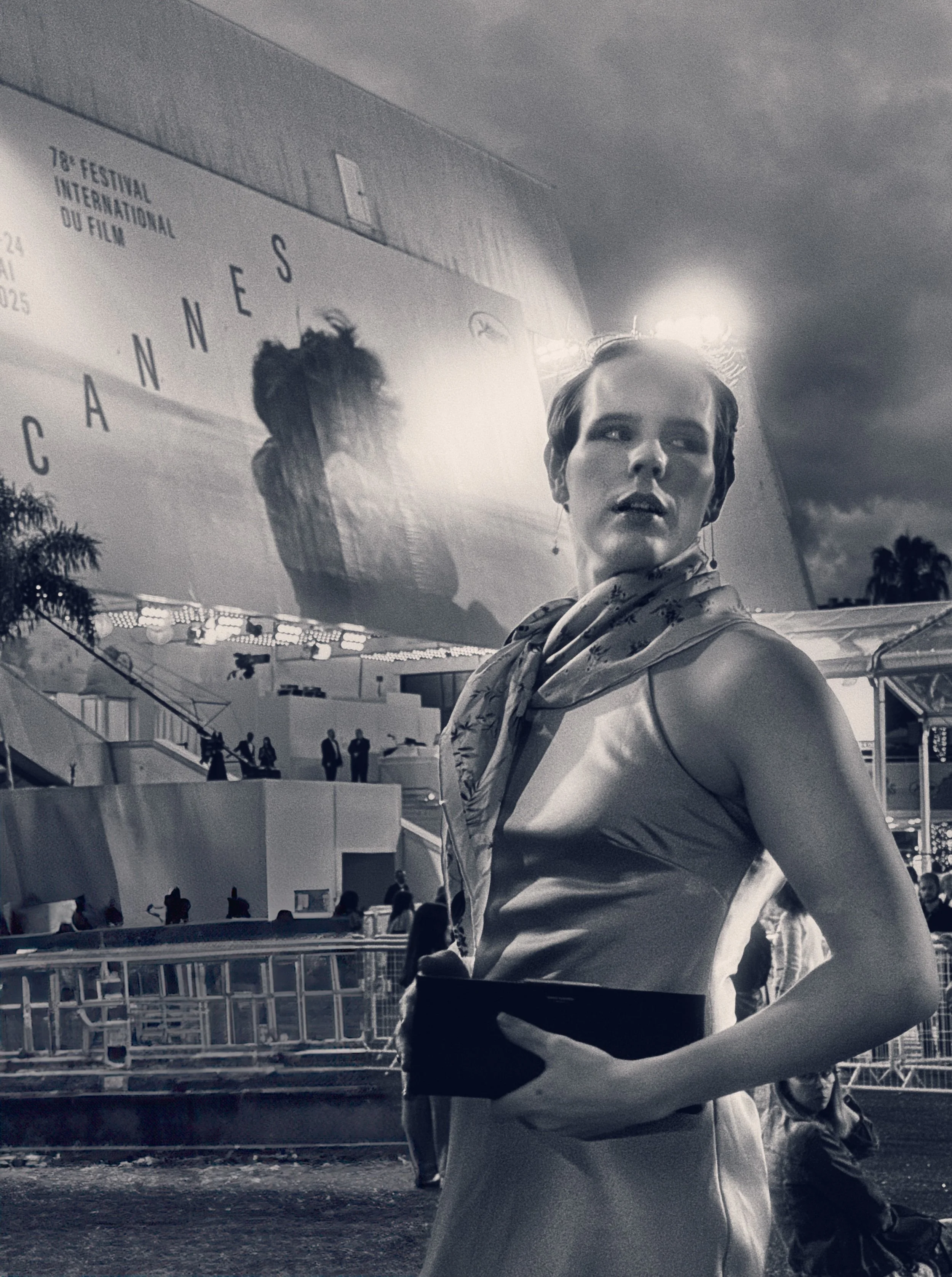 Jess Taylor in an evening gown with a scarf around her neck, standing outdoors at night near the Cannes Film Festival, with large Cannes Film Festival billboard in the background.