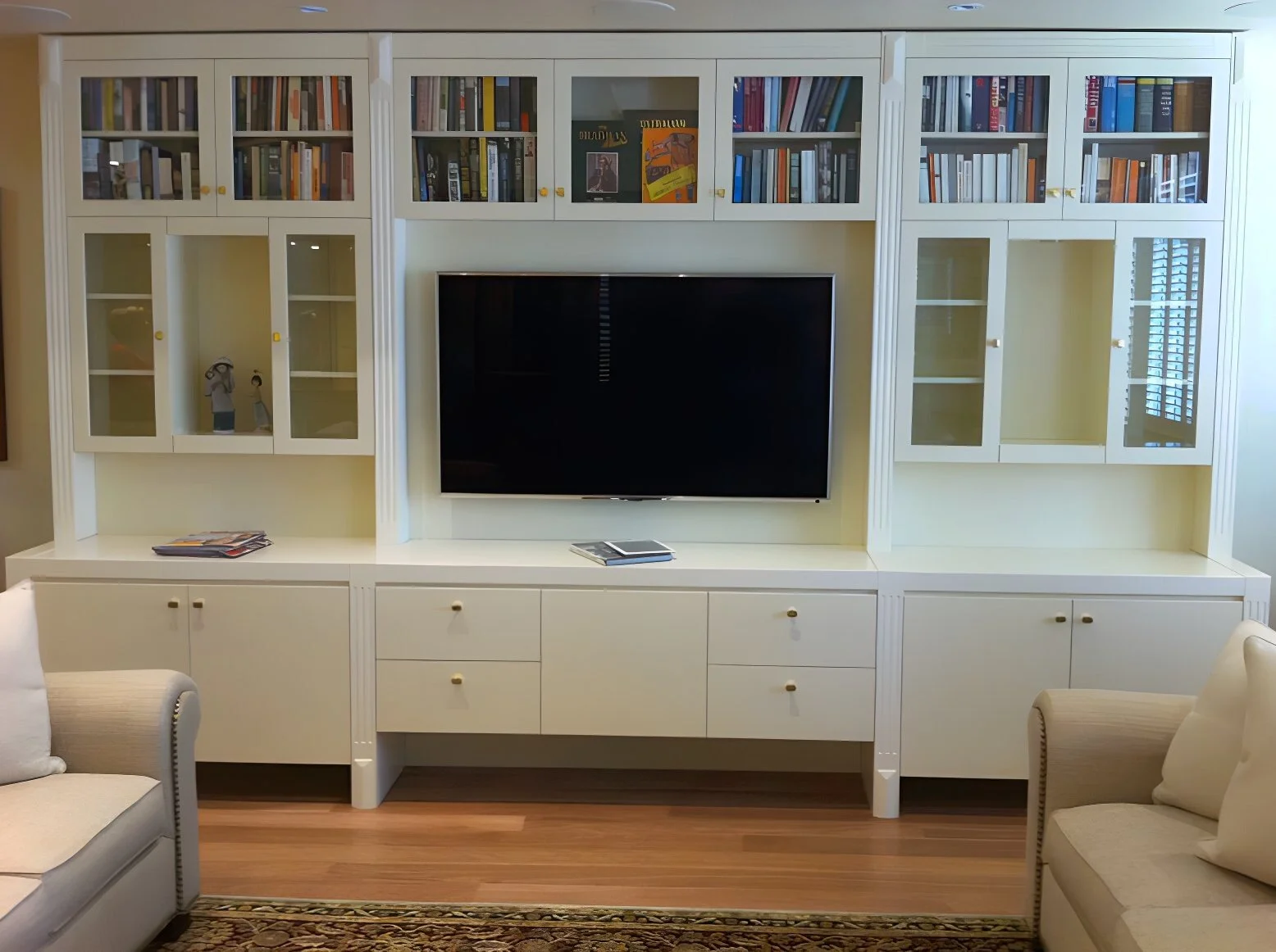 White built-in entertainment center with glass cabinets filled with books, a TV in the center, and a wood floor with a patterned rug in front.