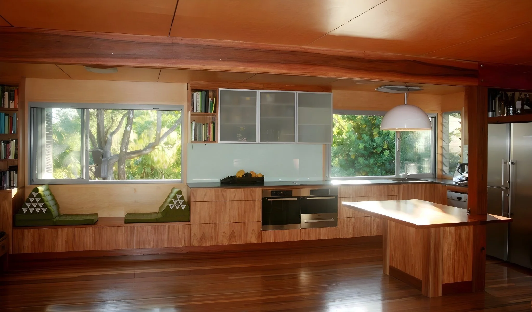 Modern kitchen with wooden cabinets, large windows showing greenery, built-in seating area, and a hanging white pendant light.