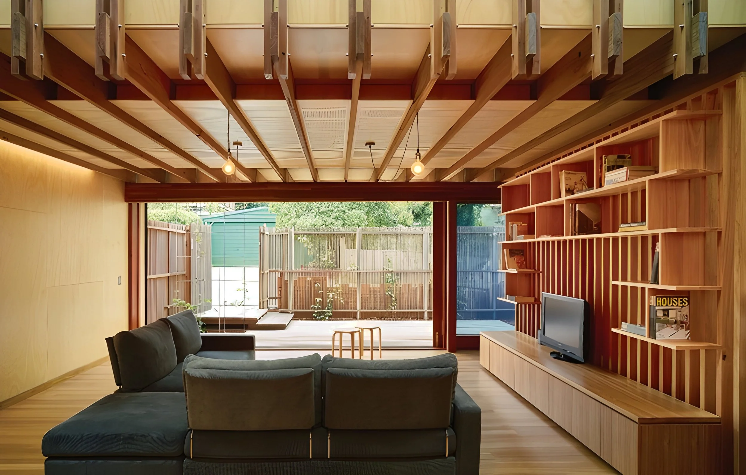 A modern living room with a black sofa, wooden shelving with books, a flat-screen TV, and large glass sliding doors leading to an outdoor garden with a wooden fence and greenery.