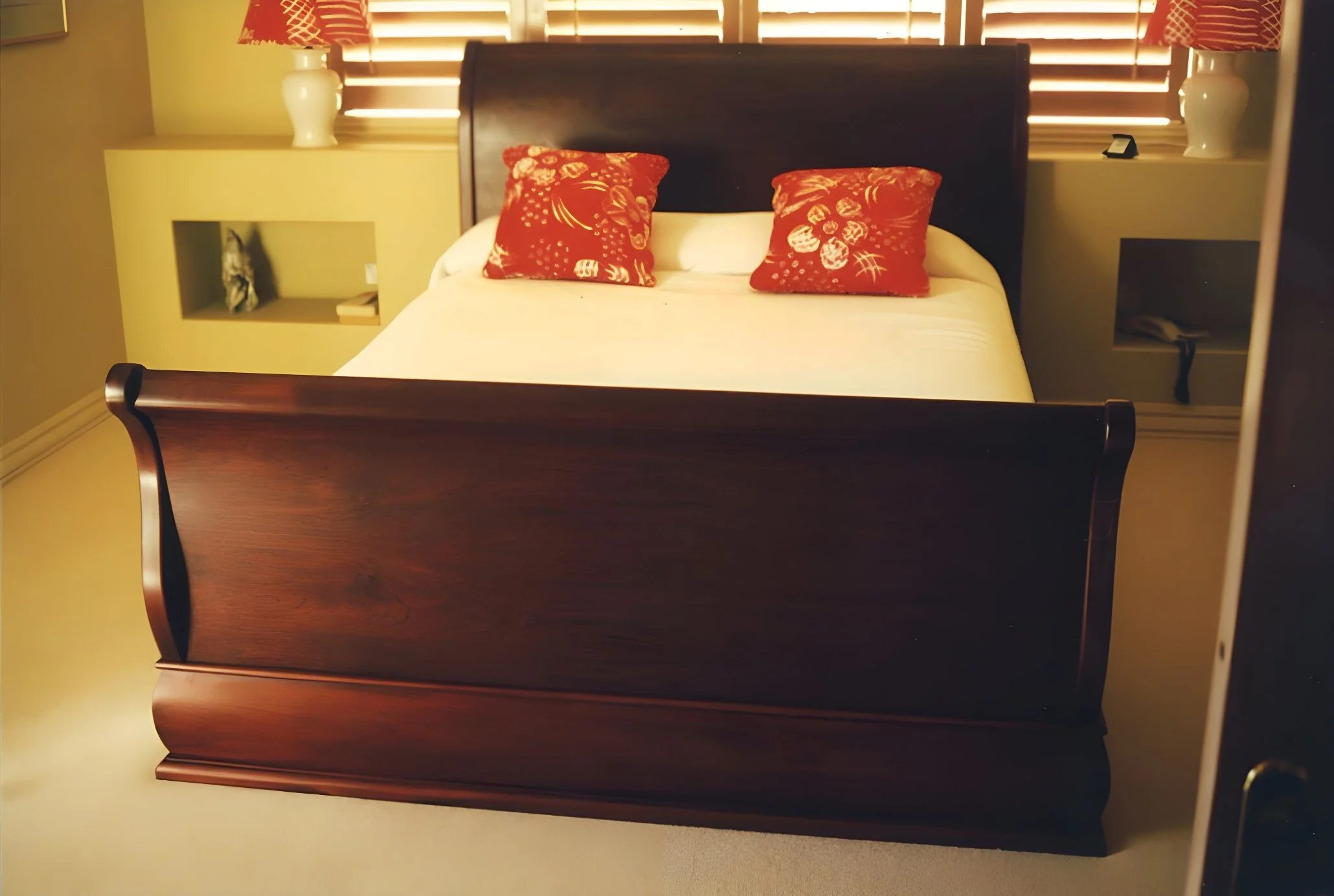 A bedroom with a wooden bed, two red decorative pillows, cream-colored sheets, a nightstand on each side with a white lamp, and window blinds behind the bed.