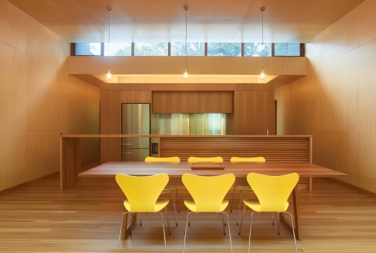 A modern kitchen and dining area with a wooden table, four yellow chairs, wooden walls, and large windows at the top letting in natural light.