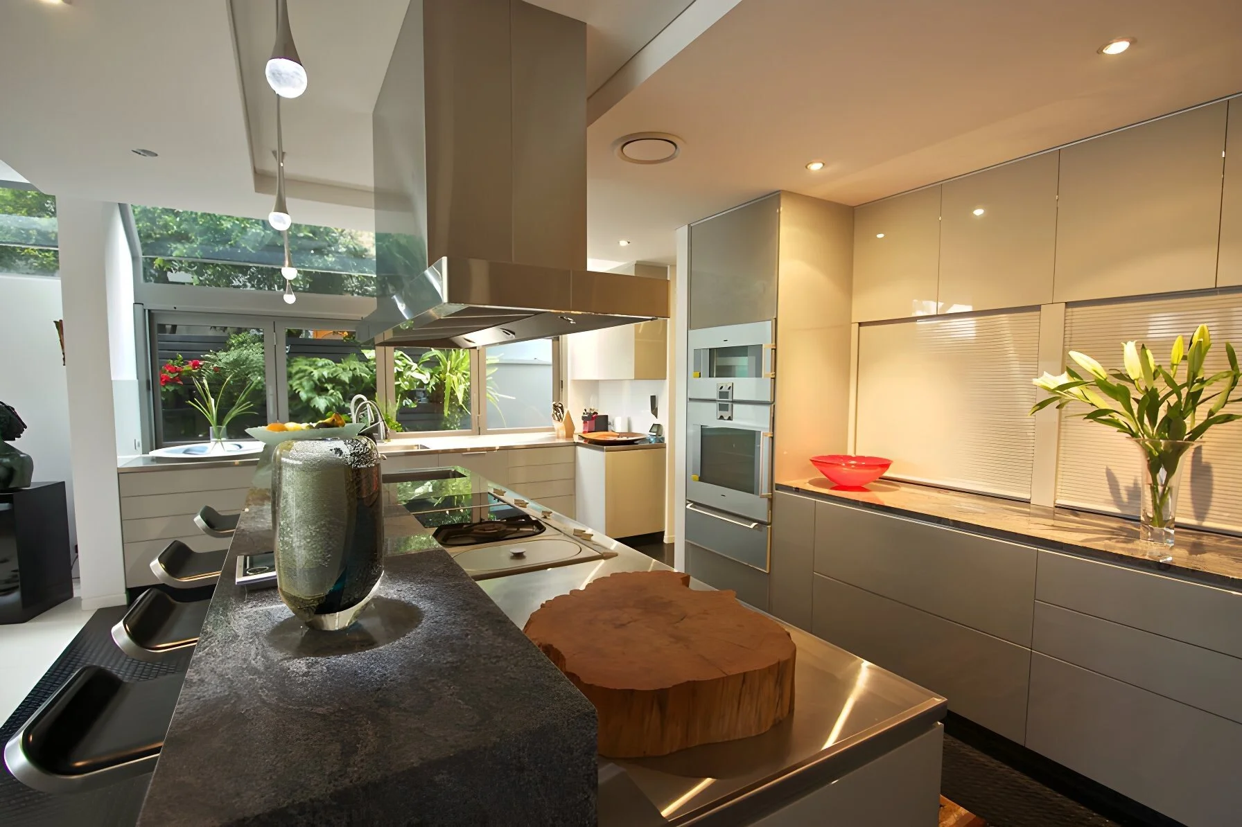 Modern kitchen with gray cabinets, marble countertops, a built-in oven, and a window overlooking greenery. Decor includes flowers, a large wooden chopping block, and a red bowl.