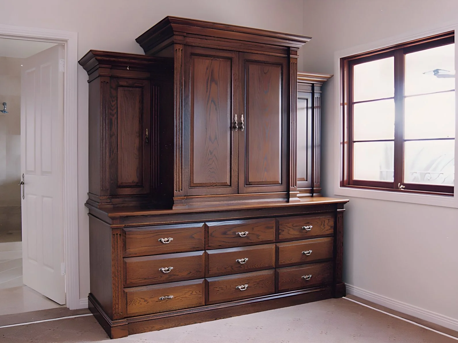 A large wooden antique-style dresser with multiple drawers and cabinets, situated next to a window with sunlight streaming in.