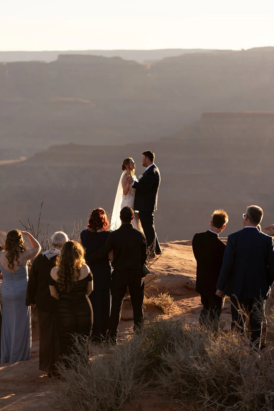 A wedding ceremony at sunset in a desert landscape, with a bride and groom standing on a rock, surrounded by guests watching as they exchange vows.