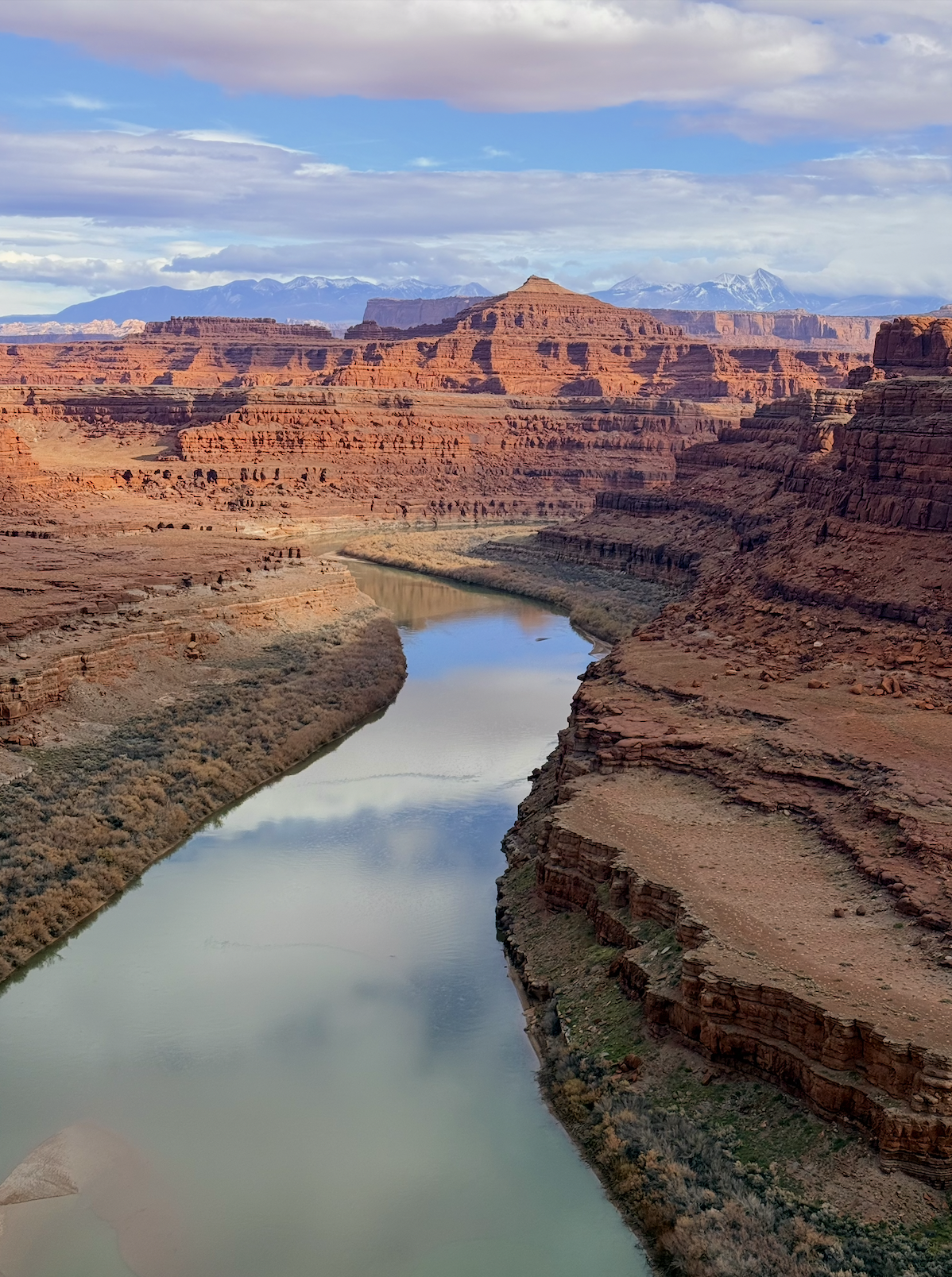 Aerial view of the Grand Canyon with a river flowing through it, red rock formations, and distant snow-capped mountains under a partly cloudy sky.