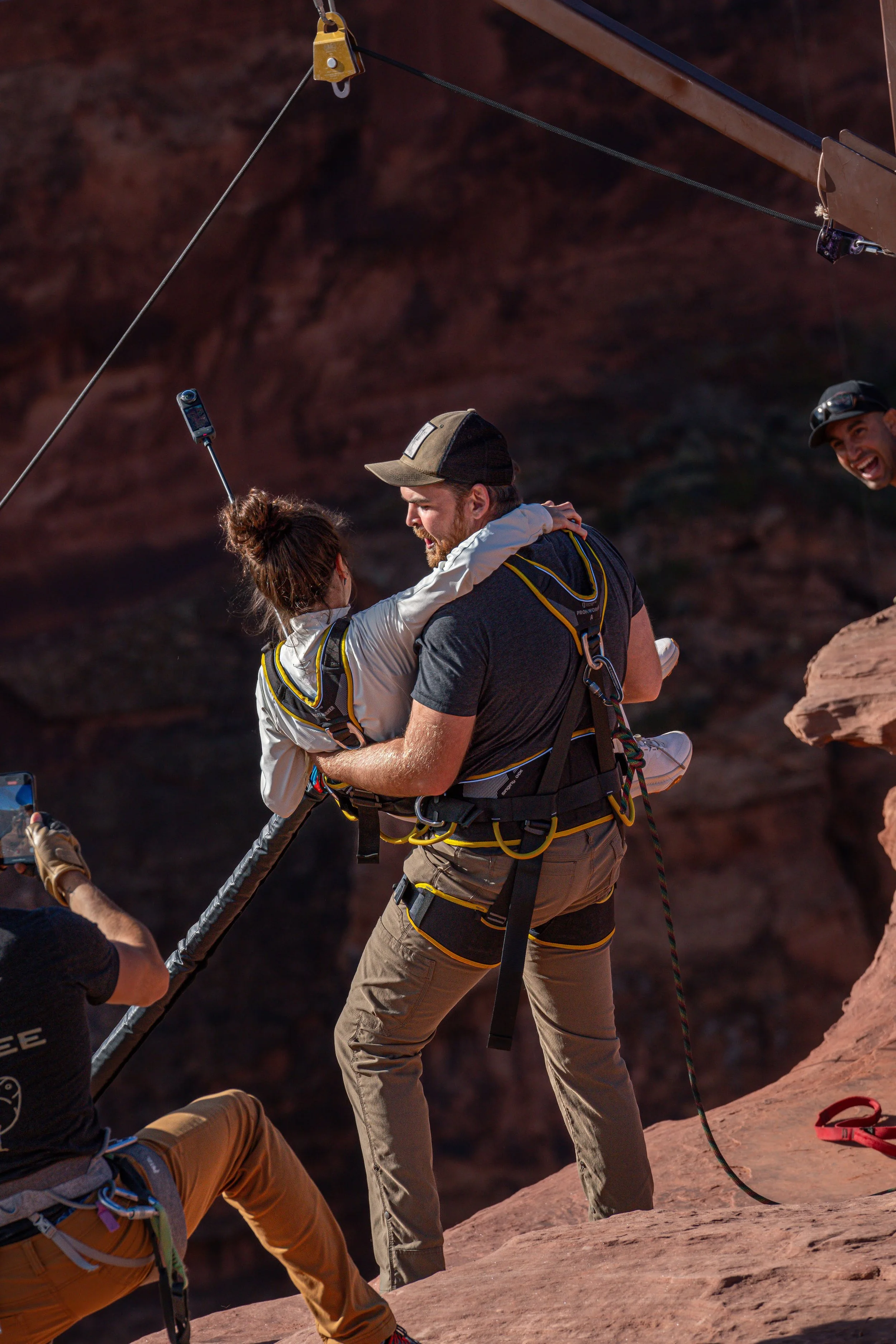 A man holding a woman in rescue gear on a rock cliff, while another man looks on and takes a photo.