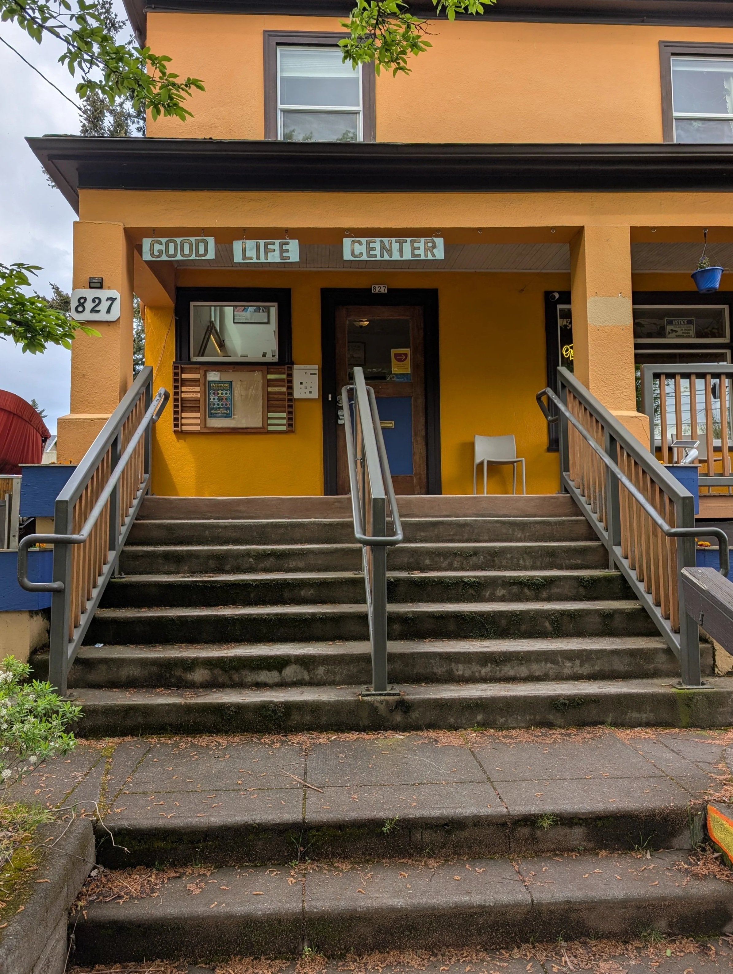 Yellow building with black accents and the sign 'Good Life Center' above the entrance, which has stairs leading up to a door, with a window and mailbox to the side, and a small chair outside.