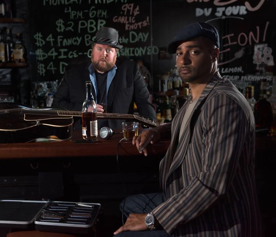 Two men at a bar, one behind and one sitting in front. The man behind has a beard, wears a fedora, a dark jacket, and a blue shirt, and is holding a glass. The man sitting has a clean haircut, wears a striped blazer and a cap, and is holding a phone. There is a guitar and a bottle on the bar counter. The background features a chalkboard with menu items written in colorful chalk.