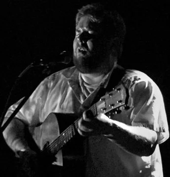 Man playing an acoustic guitar and singing into a microphone in a dark setting