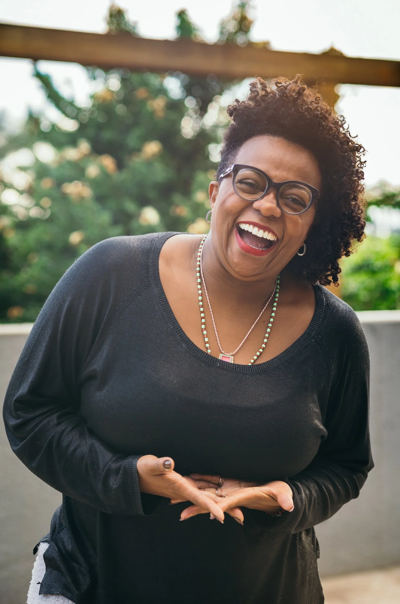 Mulher sorrindo e rindo, usando óculos, brincos e colares, com cabelo cacheado, ao ar livre com árvores ao fundo.