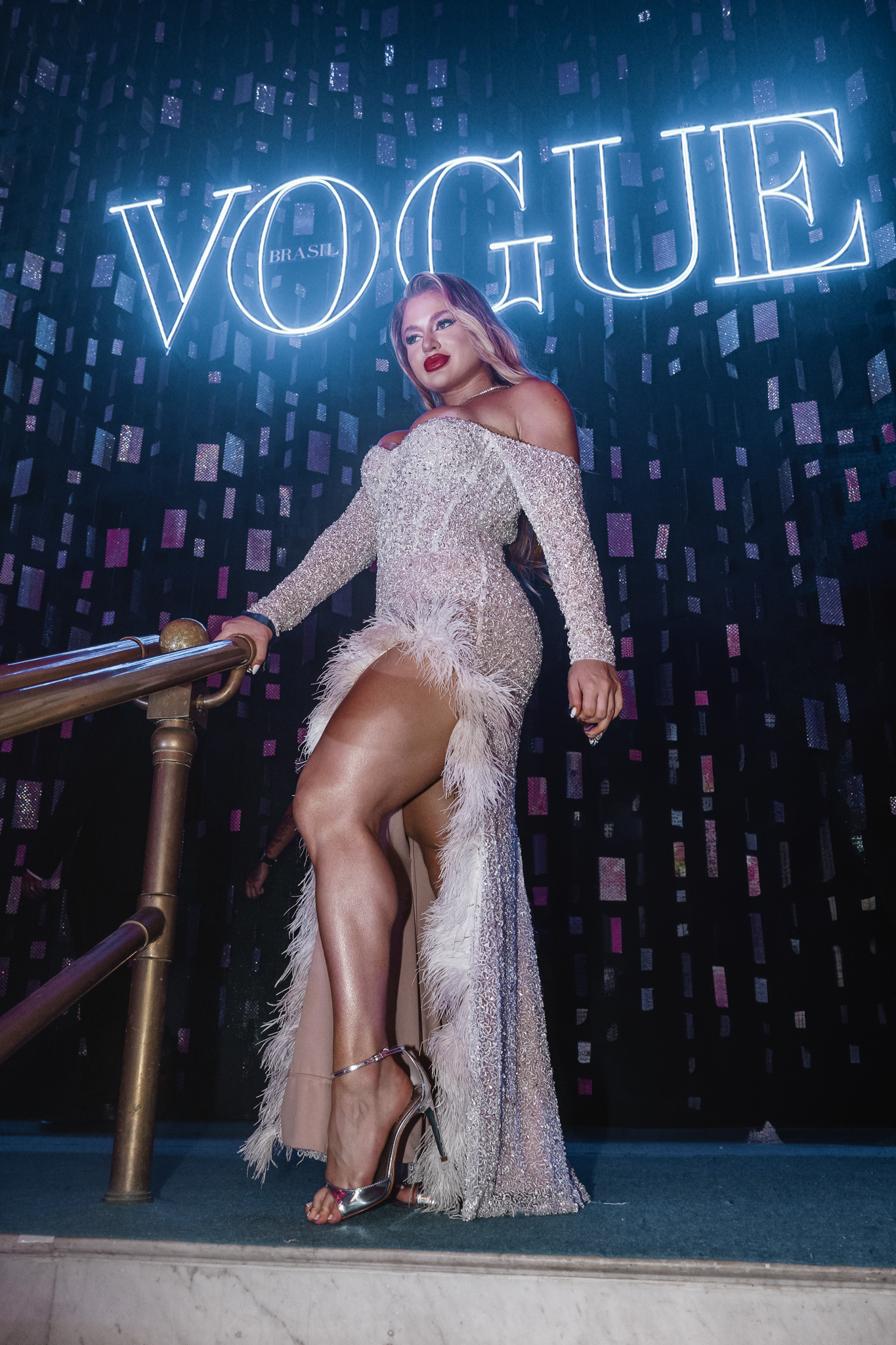 Mulher posando em frente a um letreiro de neon que diz "VOGUE BRASIL". Ela usa um vestido brilhante com detalhes de pena na parte inferior, com brilho e glamour.