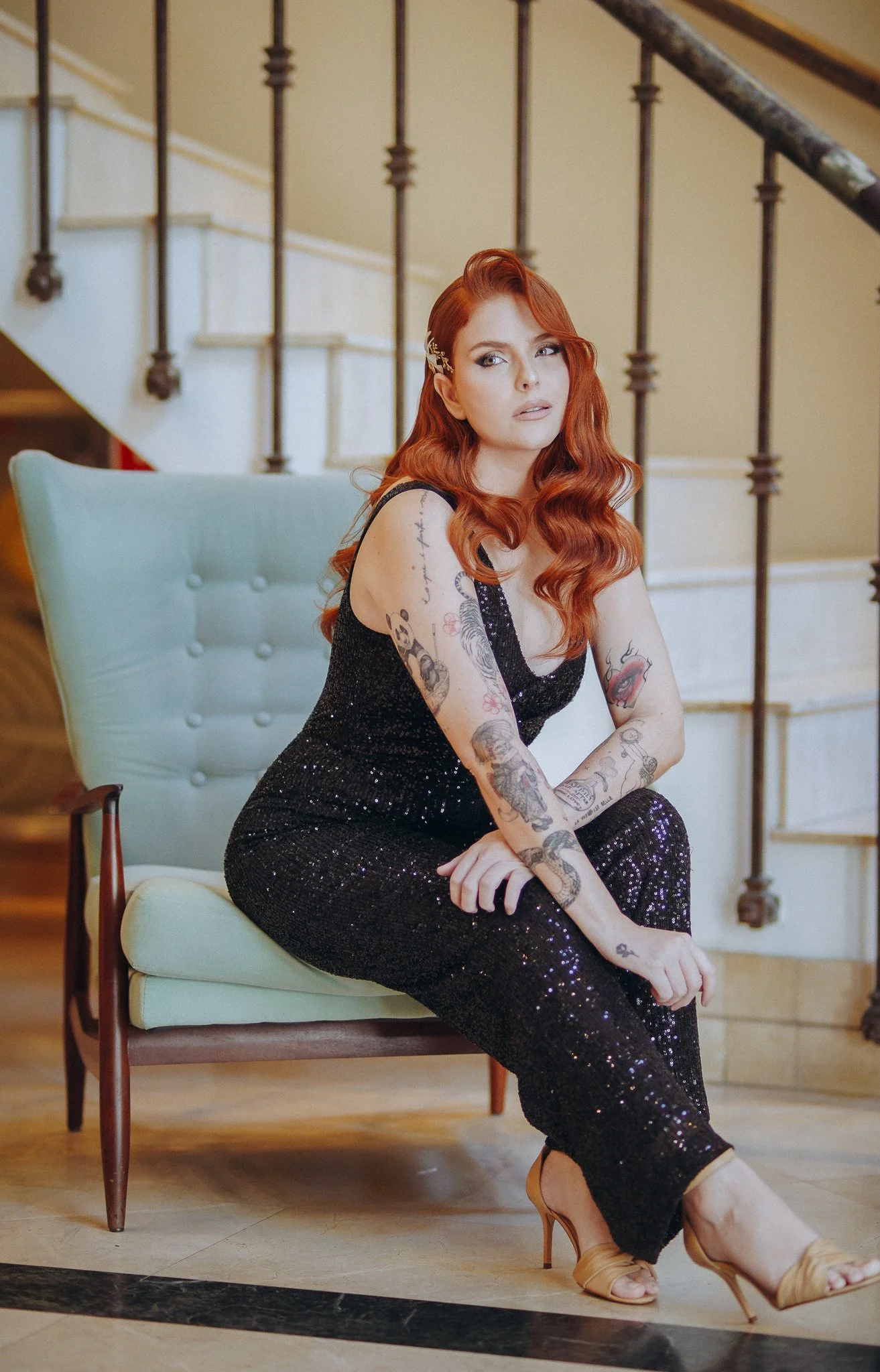 Mulher com cabelo ruivo e tatuagens, vestindo vestido preto com brilhos, sentada em uma poltrona suave, em um ambiente com escada de ferro ao fundo.