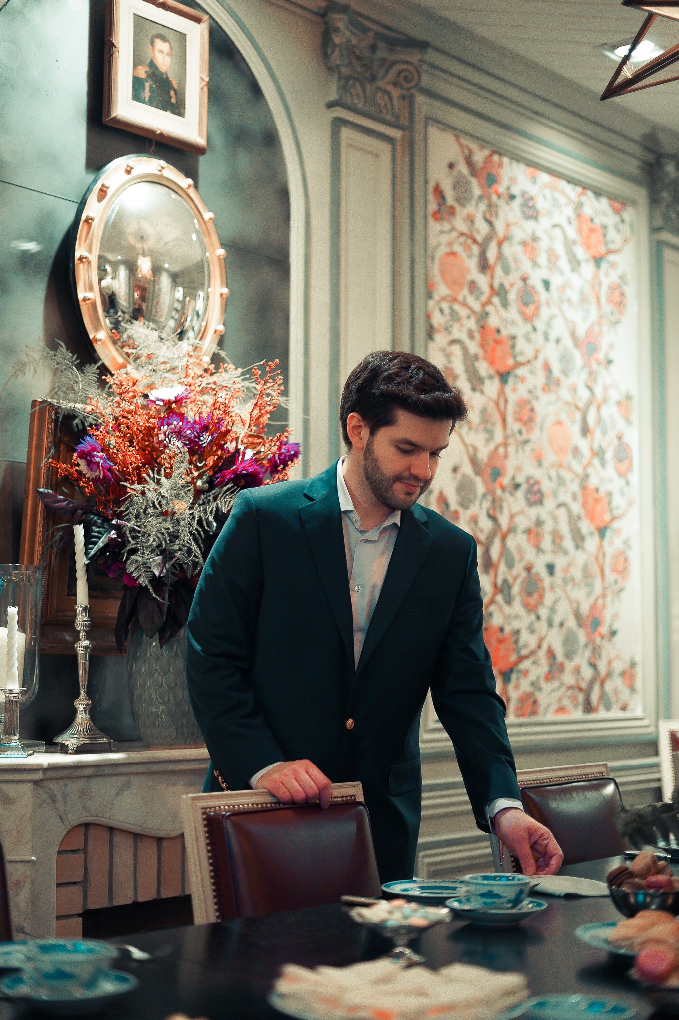Homem de terno organizando uma mesa de jantar em um ambiente decorado com arranjos de flores e velas.