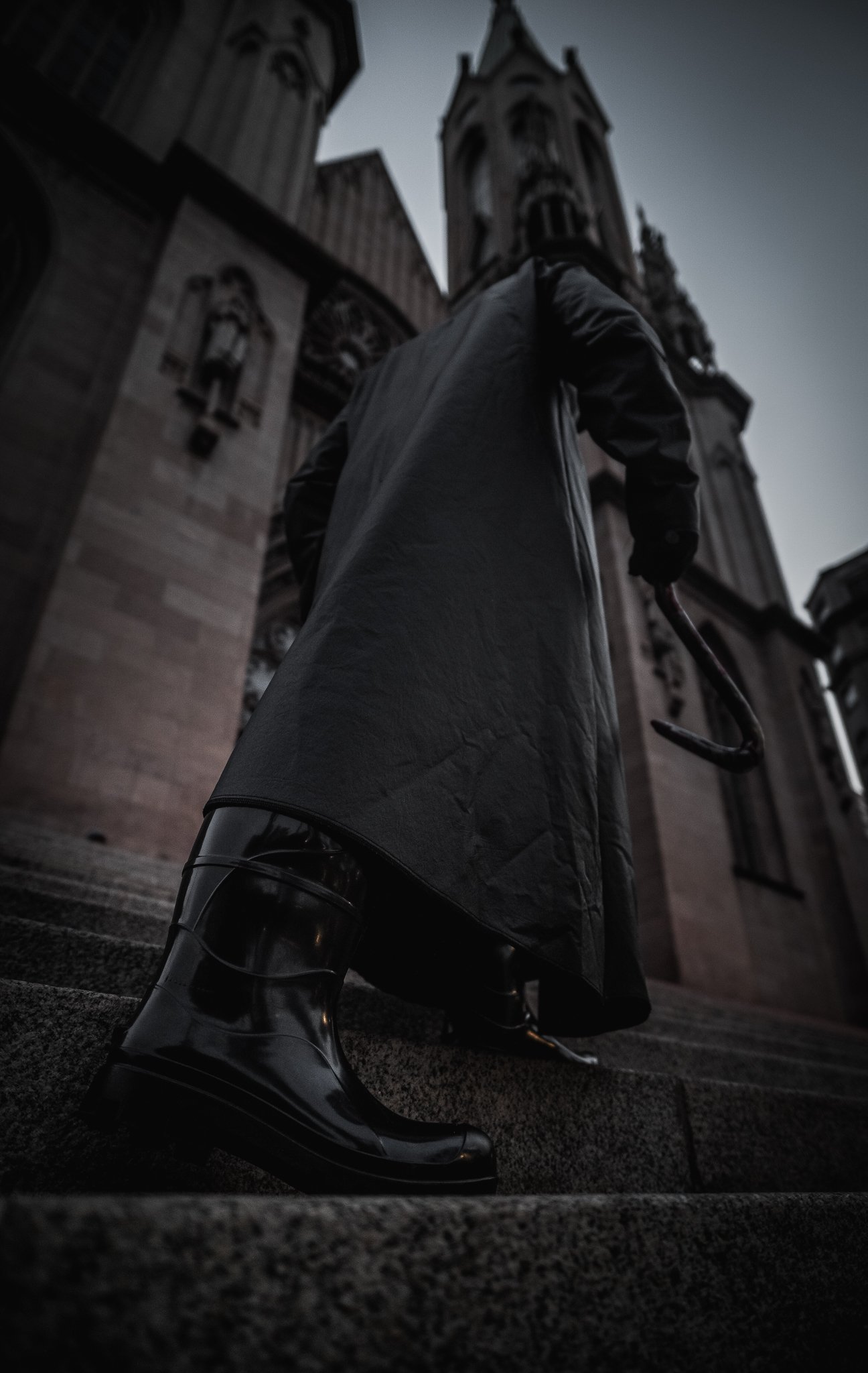 Pessoa de costas segurando um guarda-chuva na frente de uma igreja gótica com torres altas e janelas arqueadas ao entardecer.