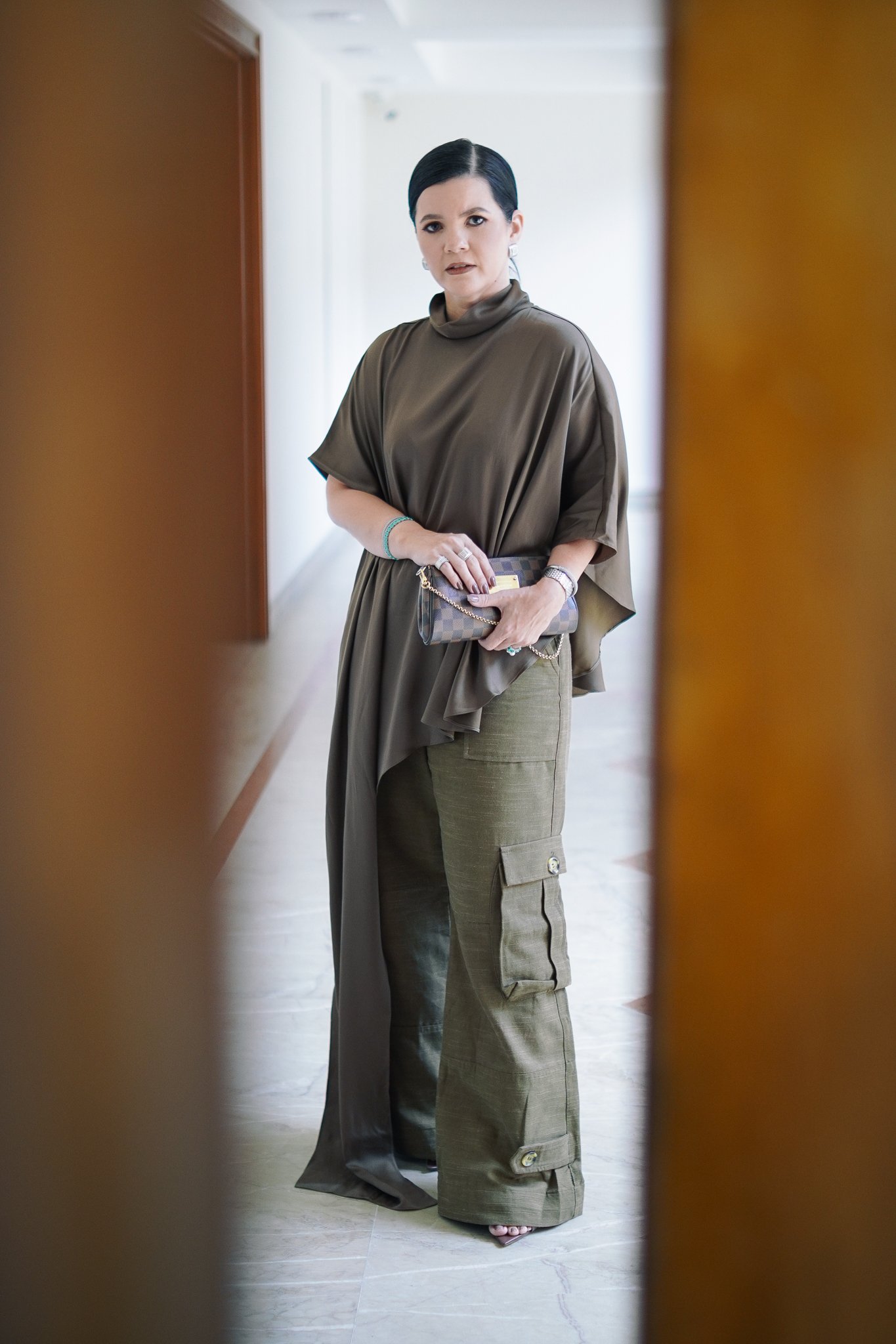 Mulher com cabelo escuro e roupa de tom oliva, segurando uma bolsa de mão pequena com detalhes dourados, olhando para a câmera enquanto está na passagem de um corredor, com paredes brancas ao fundo.