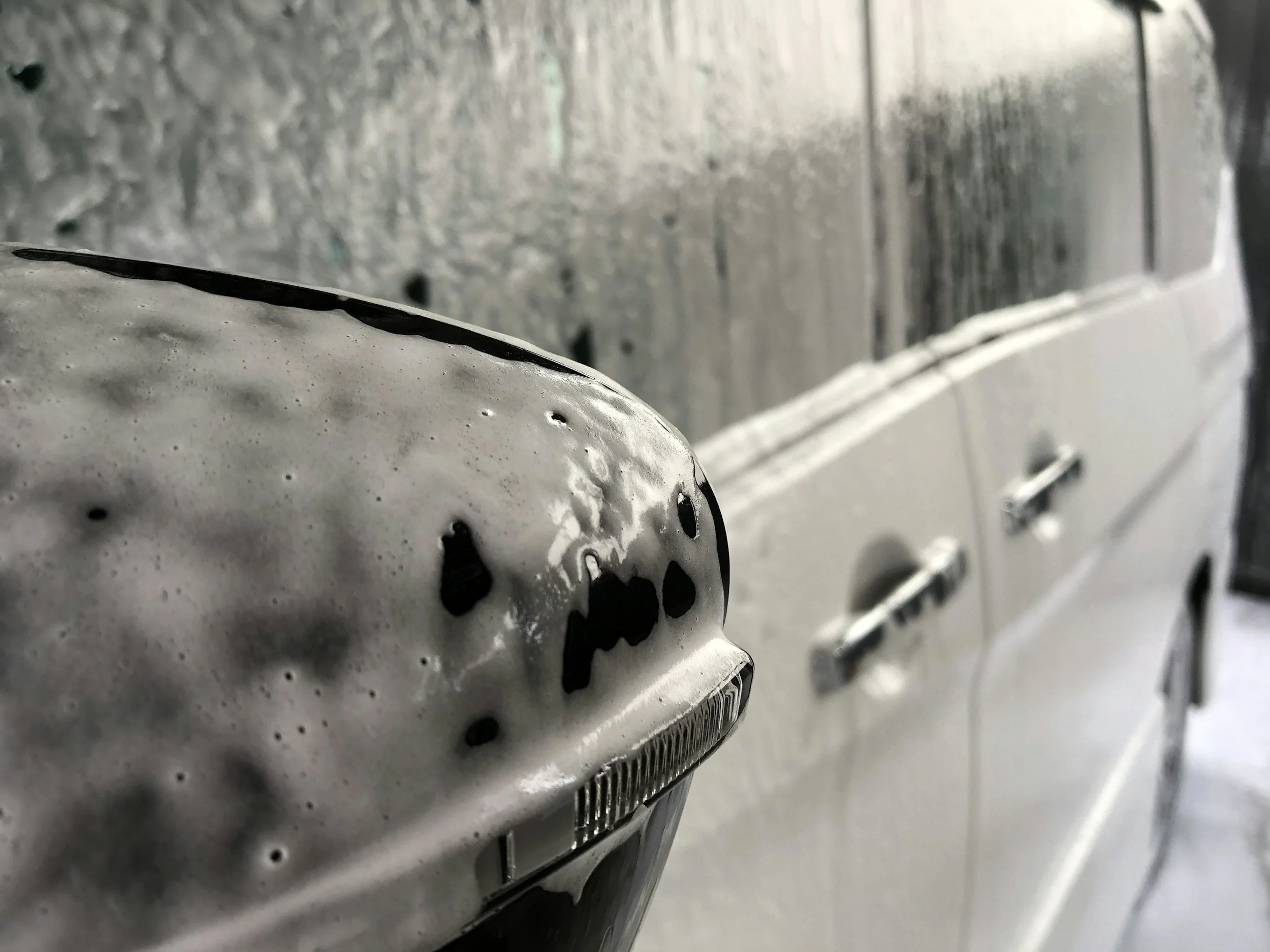 Close-up of a white car's wing mirror and side, covered in soap suds, snow foam.