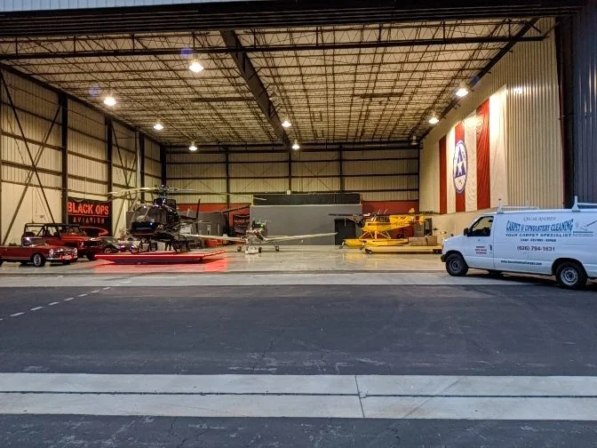 Indoor aircraft hangar with helicopters and planes, including a black helicopter, a red aircraft, and a yellow seaplane, along with a white service van and various hangar equipment.