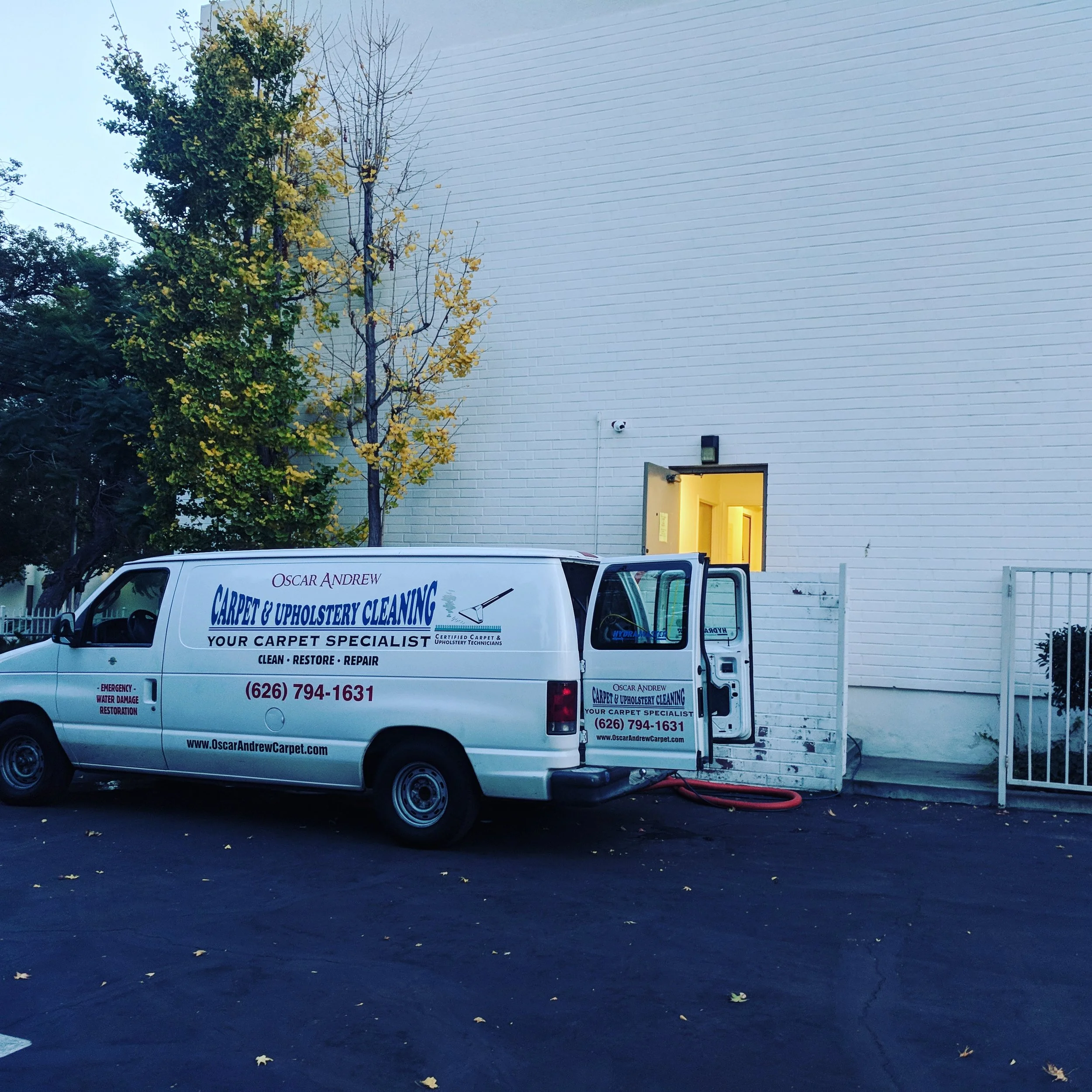 A white van parked outside a building with an open door, with a hose connected to the back for carpet cleaning. The van displays contact information and services, including carpet and upholstery cleaning.