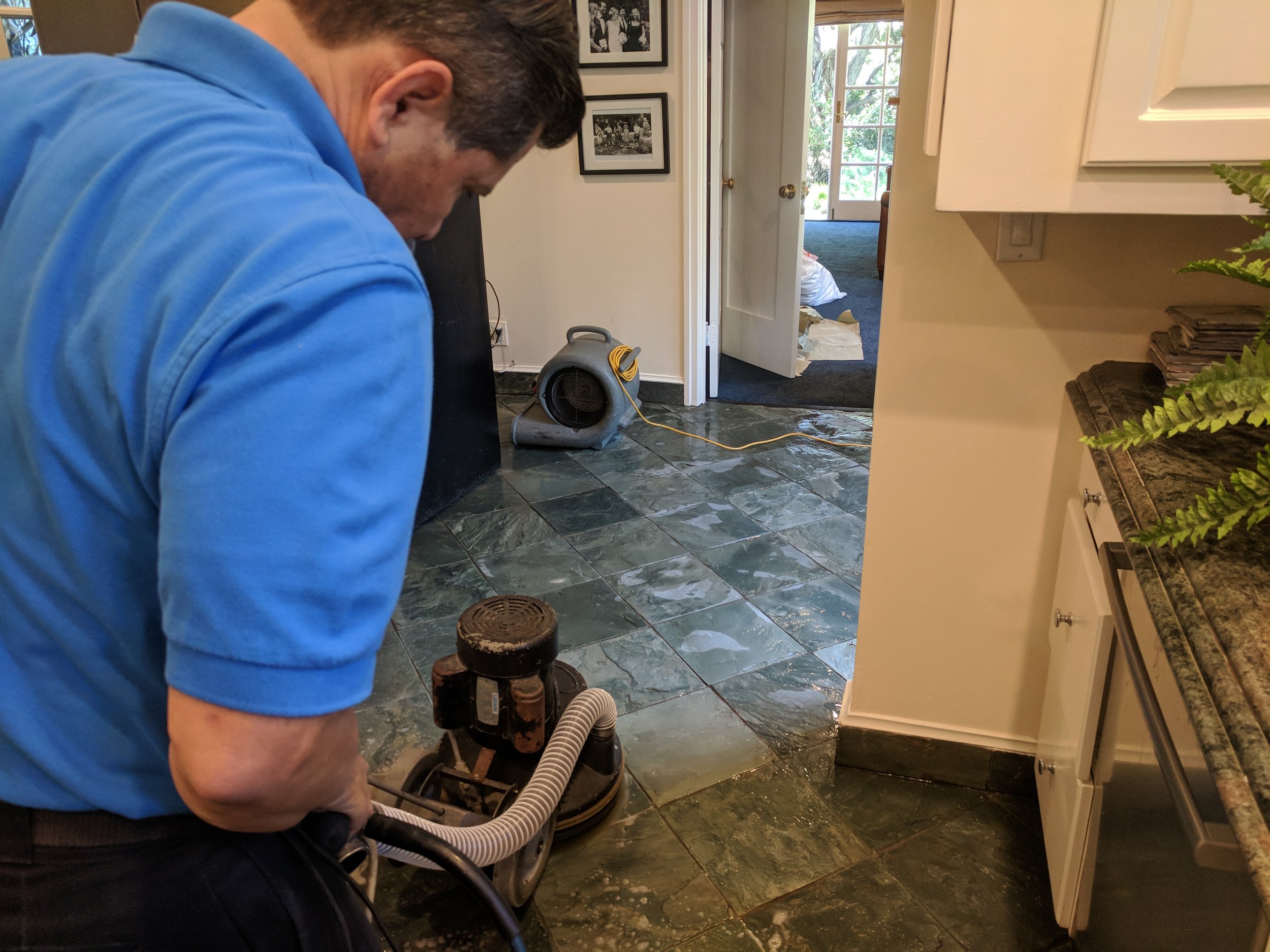 A man in a blue shirt using a wet vacuum cleaner on a stone kitchen floor.