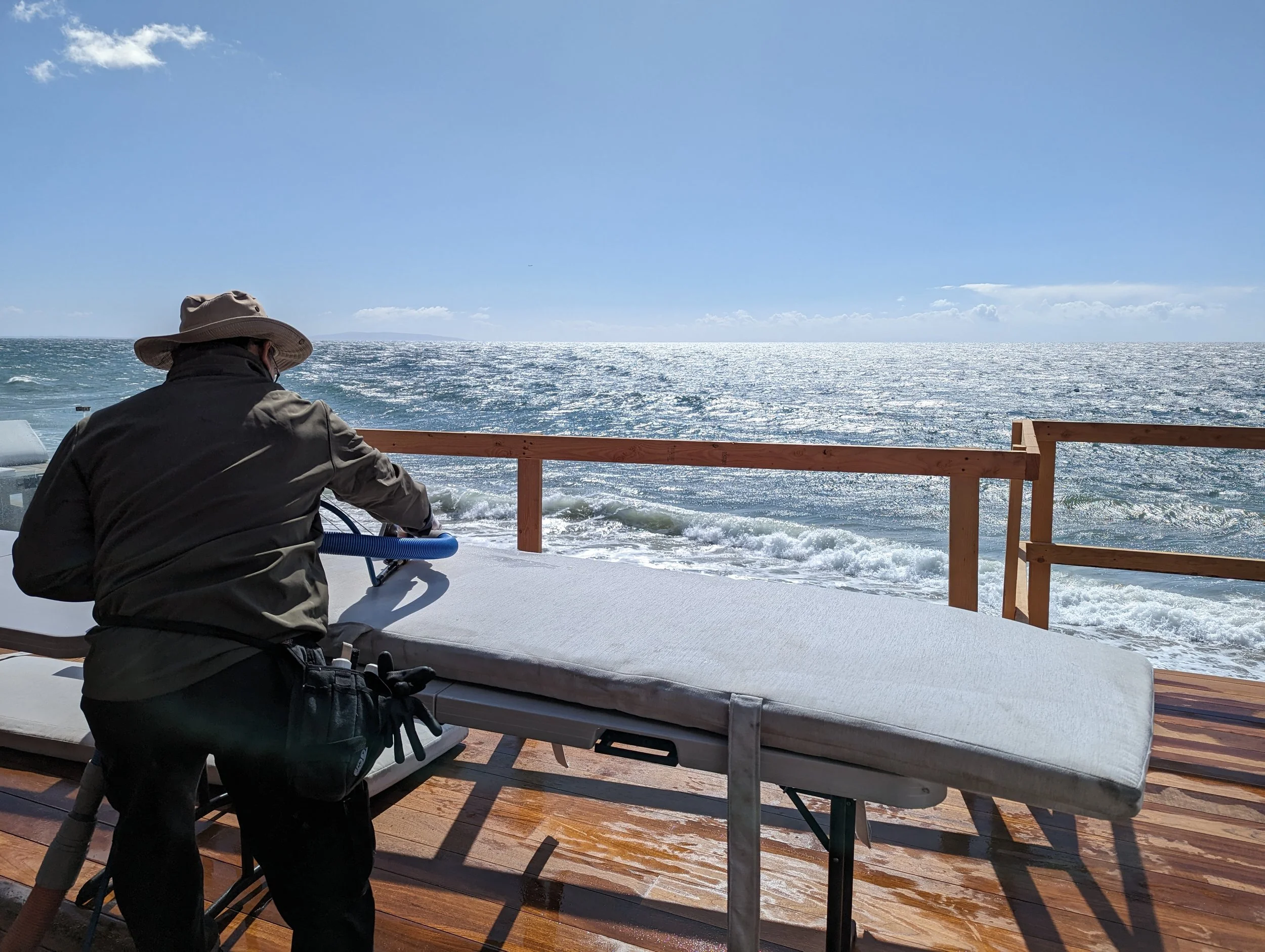 A person wearing a hat and jacket setting up or adjusting a massage table on a wooden deck near the ocean, with the sea and sky in the background.