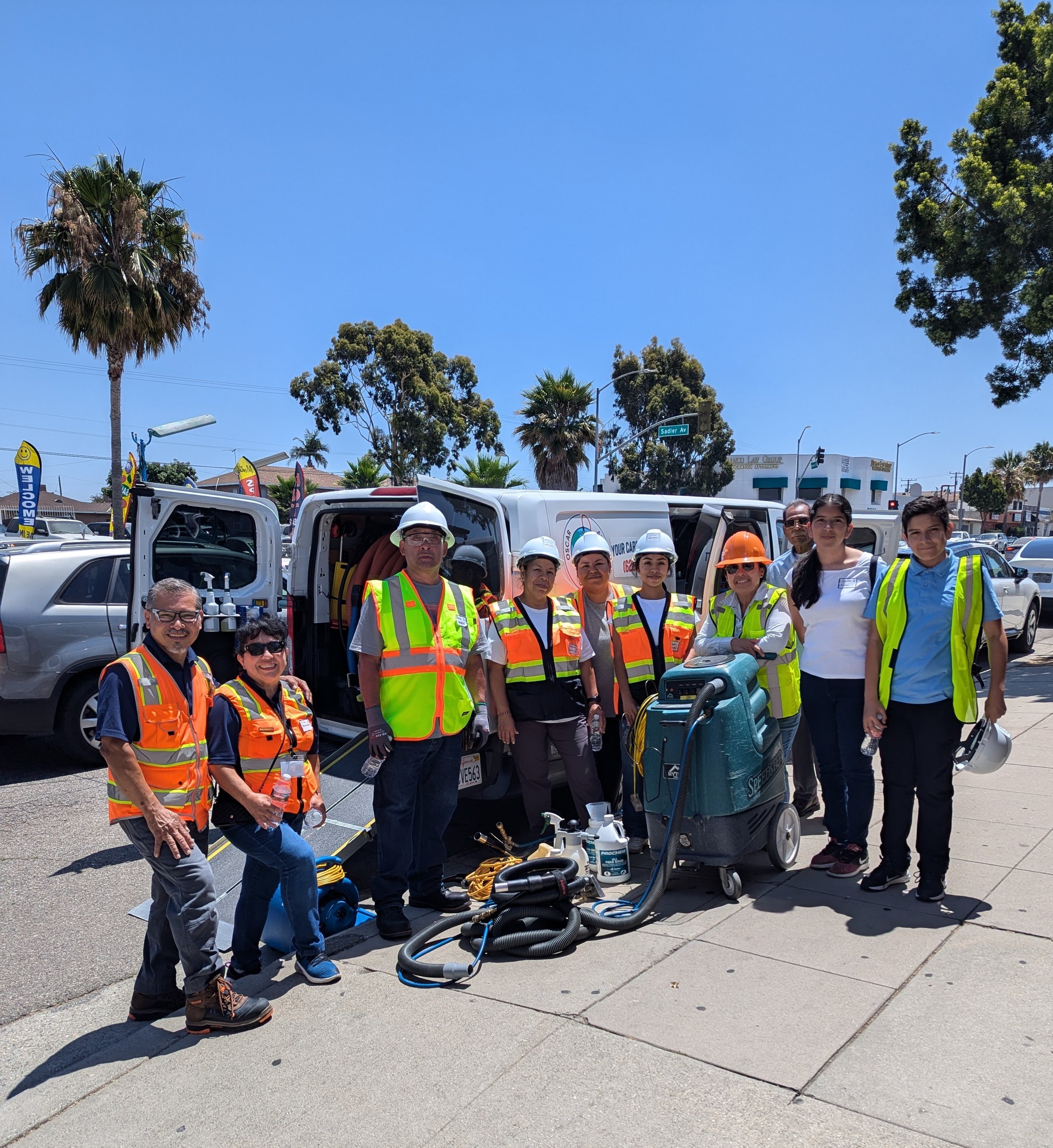 Group of workers in safety vests and helmets standing in front of a van with equipment, on a sunny day, in an urban area.