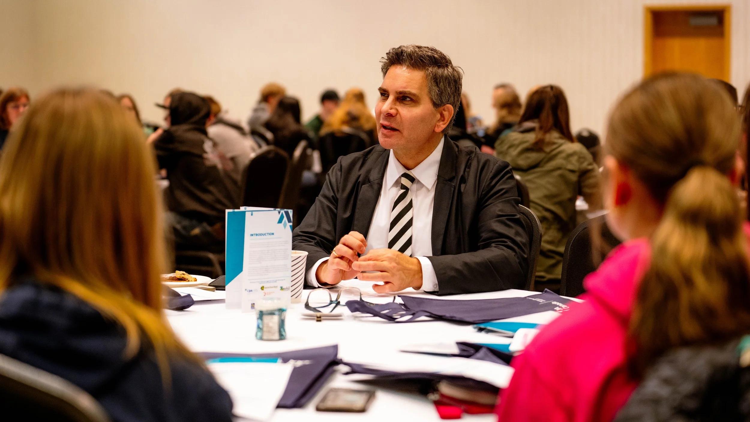 A man in a suit and striped tie engaged in conversation at a roundtable during a conference or event with a group of women around him, in a large conference room.