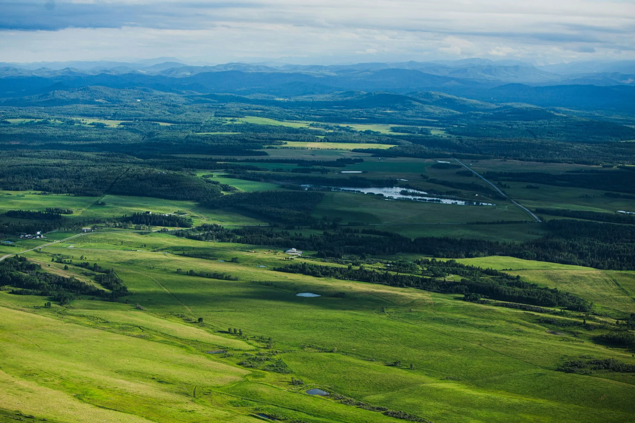 Aerial view of lush green fields, forests, and small bodies of water in a rural landscape with mountains in the background under cloudy skies.