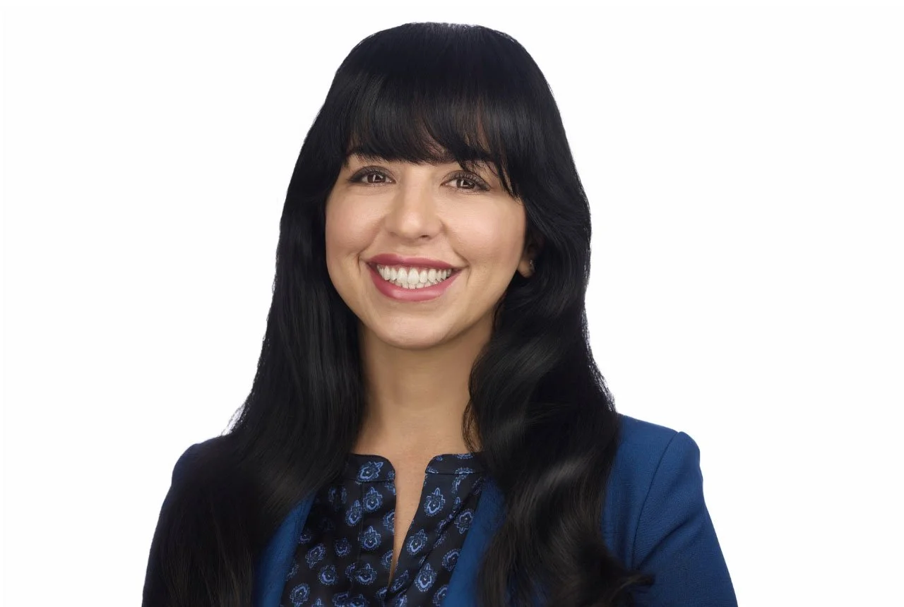 A woman with long black hair and a bright smile, wearing a blue blazer and a patterned blouse, against a white background.