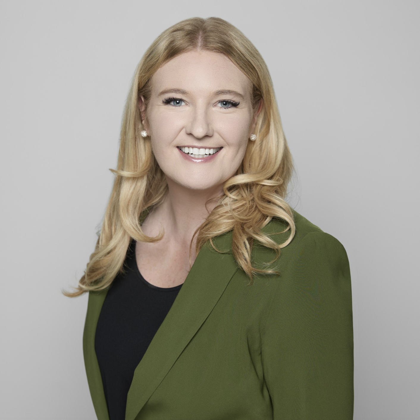 Headshot of a woman with blonde hair, wearing a green blazer and a black top, smiling against a plain grey background.