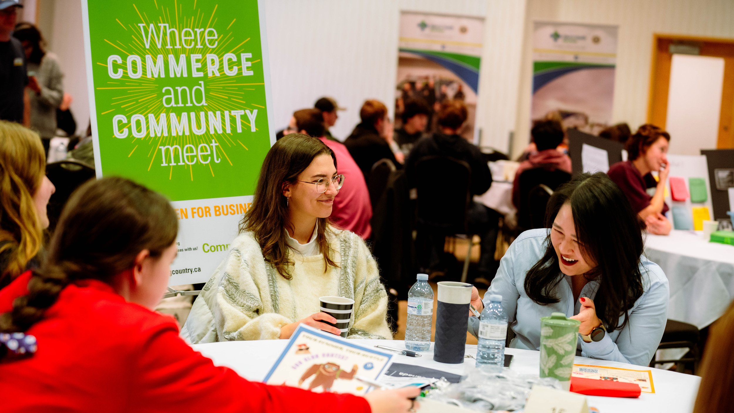 A group of people sitting around a table at a community meet, with a green sign that reads 'Where Commerce and Community Meet' in the background.
