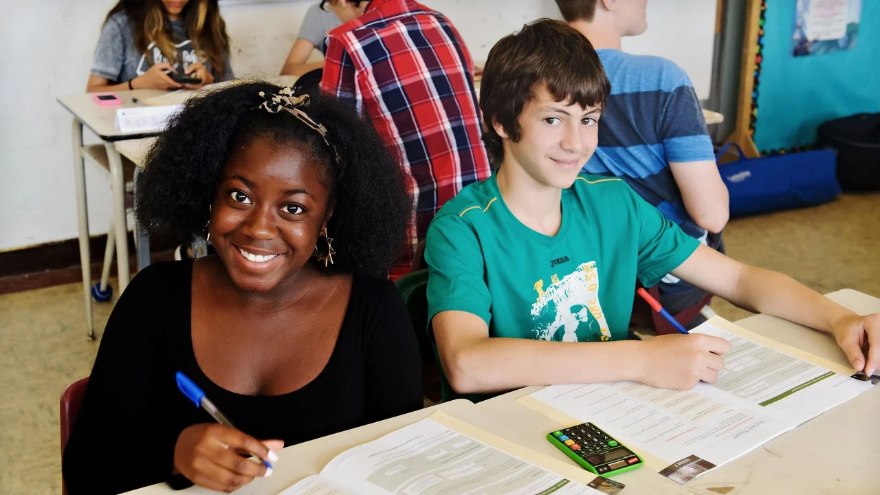 A classroom with students sitting at desks, focusing on their work. In the foreground, a smiling girl with dark, curly hair and wearing a black shirt holds a pen. Next to her, a boy with brown hair and a green shirt also works with a pen. The background shows other students and classroom materials.