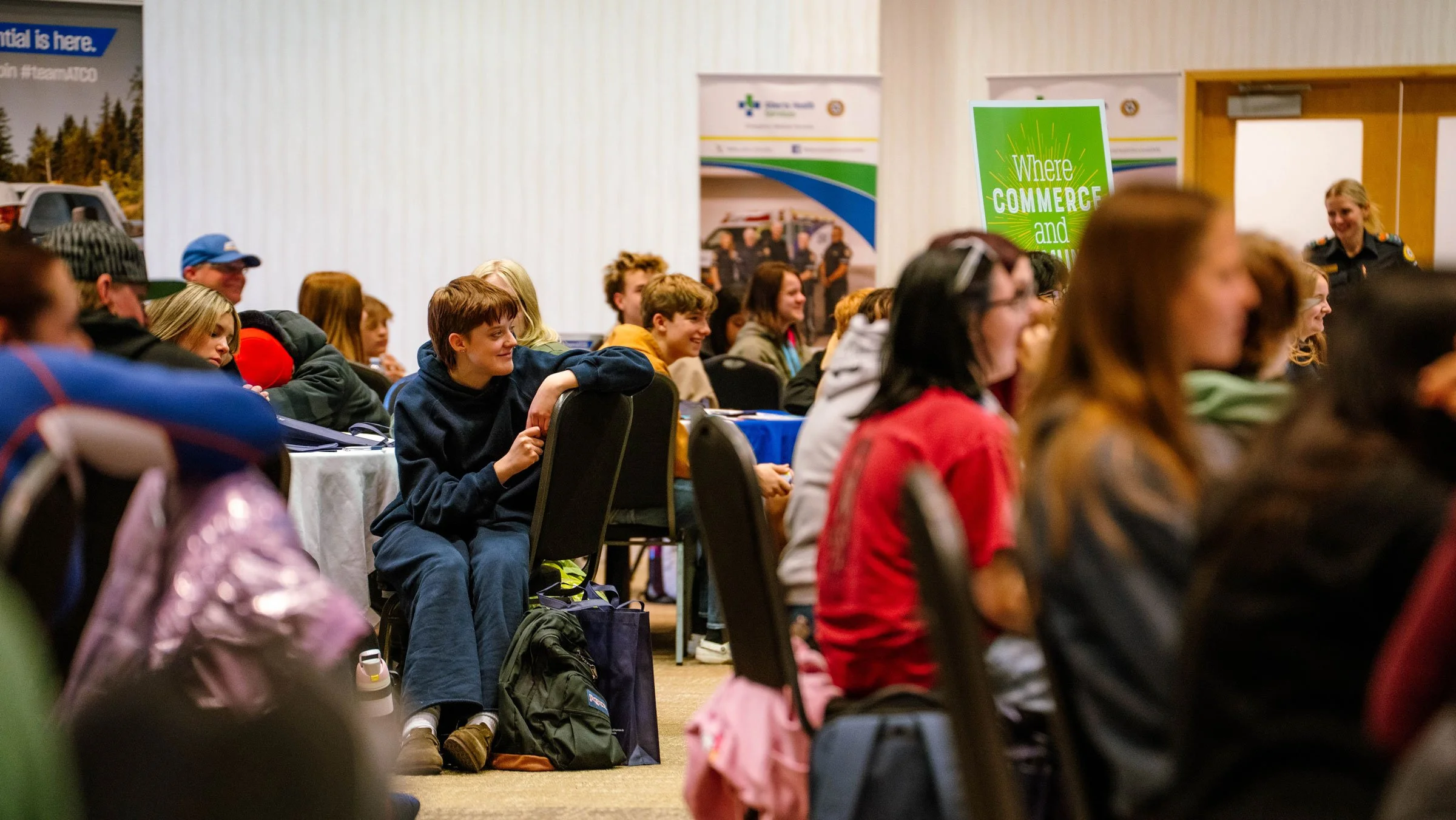 A group of young people sitting in a conference room, attending an event or presentation, with a few booths and banners visible in the background.