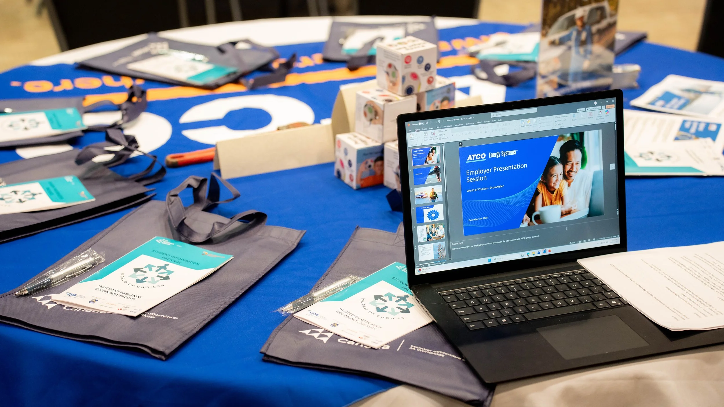 Table with ATCO Energy Systems promotional materials, tote bags, pens, and a laptop displaying a presentation titled 'Employer Presentation Session' with a family on the slide.