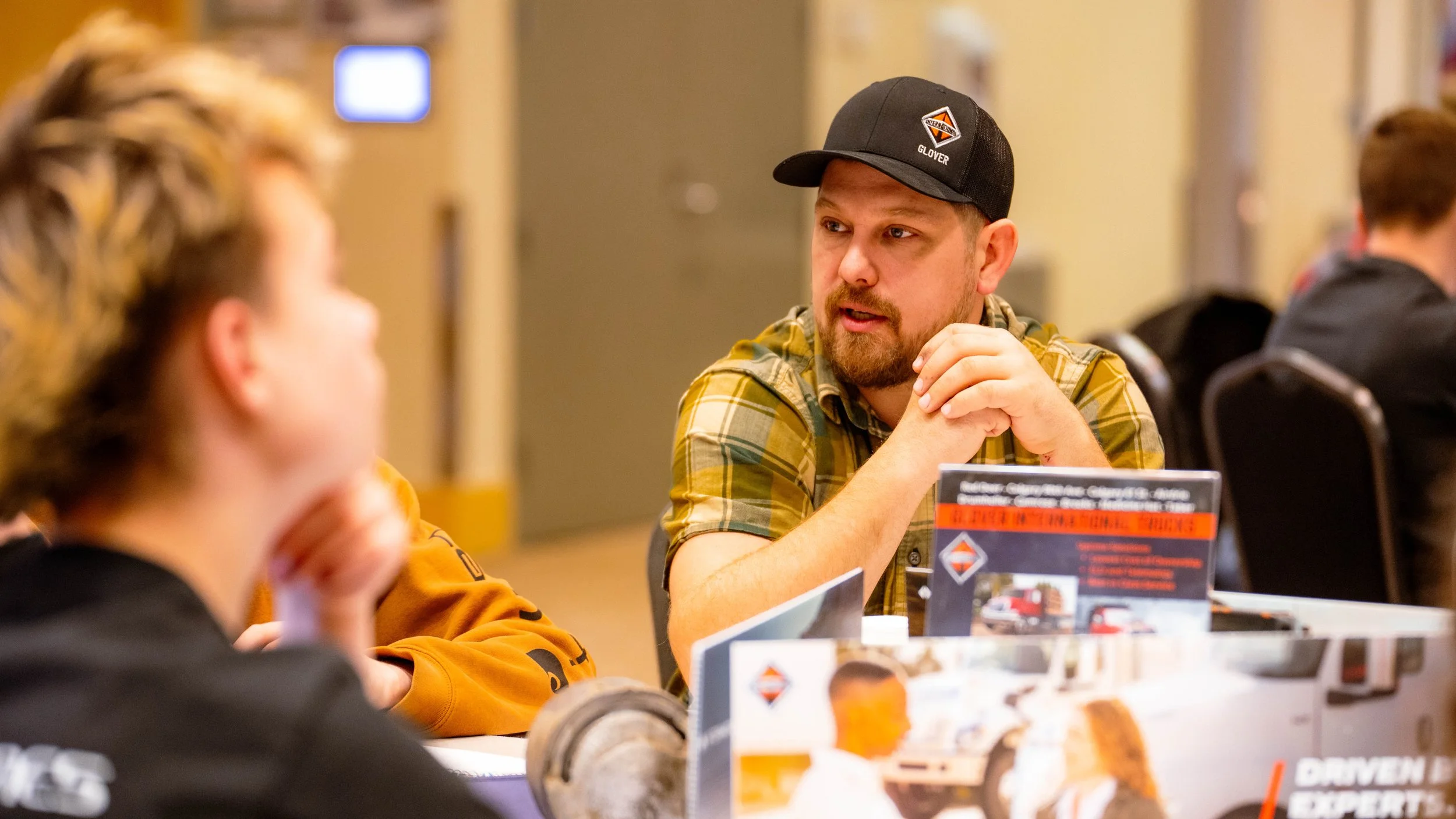 A man wearing a black trucker hat and a green plaid shirt is sitting at a table, engaged in conversation with a woman who has curly hair, wearing an orange shirt. The scene appears to be in a public setting, possibly a conference or meeting, with informational flyers and banners on the table.