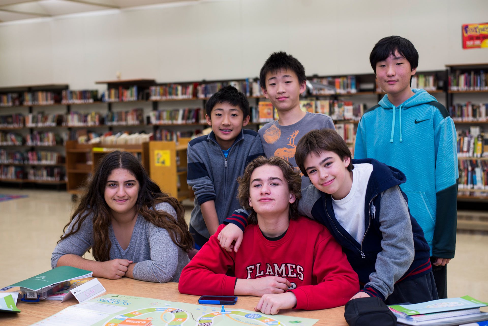 Group of six diverse young students in a school library, gathered around a decorated table with books and papers, smiling for the camera.