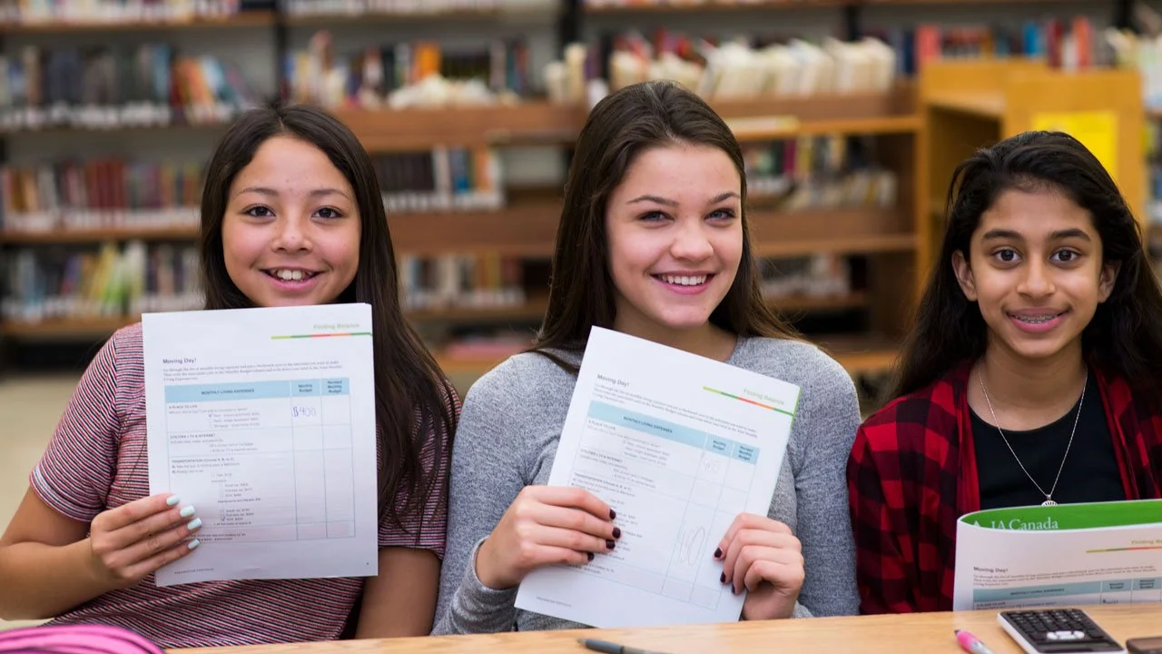 Three students smiling and holding report cards while seated at a table in a library during a JA (Junior Achievement) Southern Alberta program