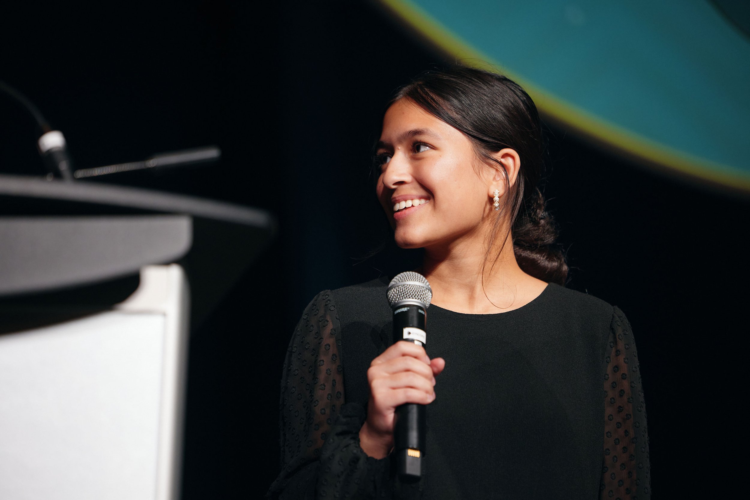 A woman with dark hair in a braid, wearing pearl earrings and a black dress with sheer sleeves, smiling while holding a microphone, standing near a keyboard on a stage with a dark background and green and yellow lights.