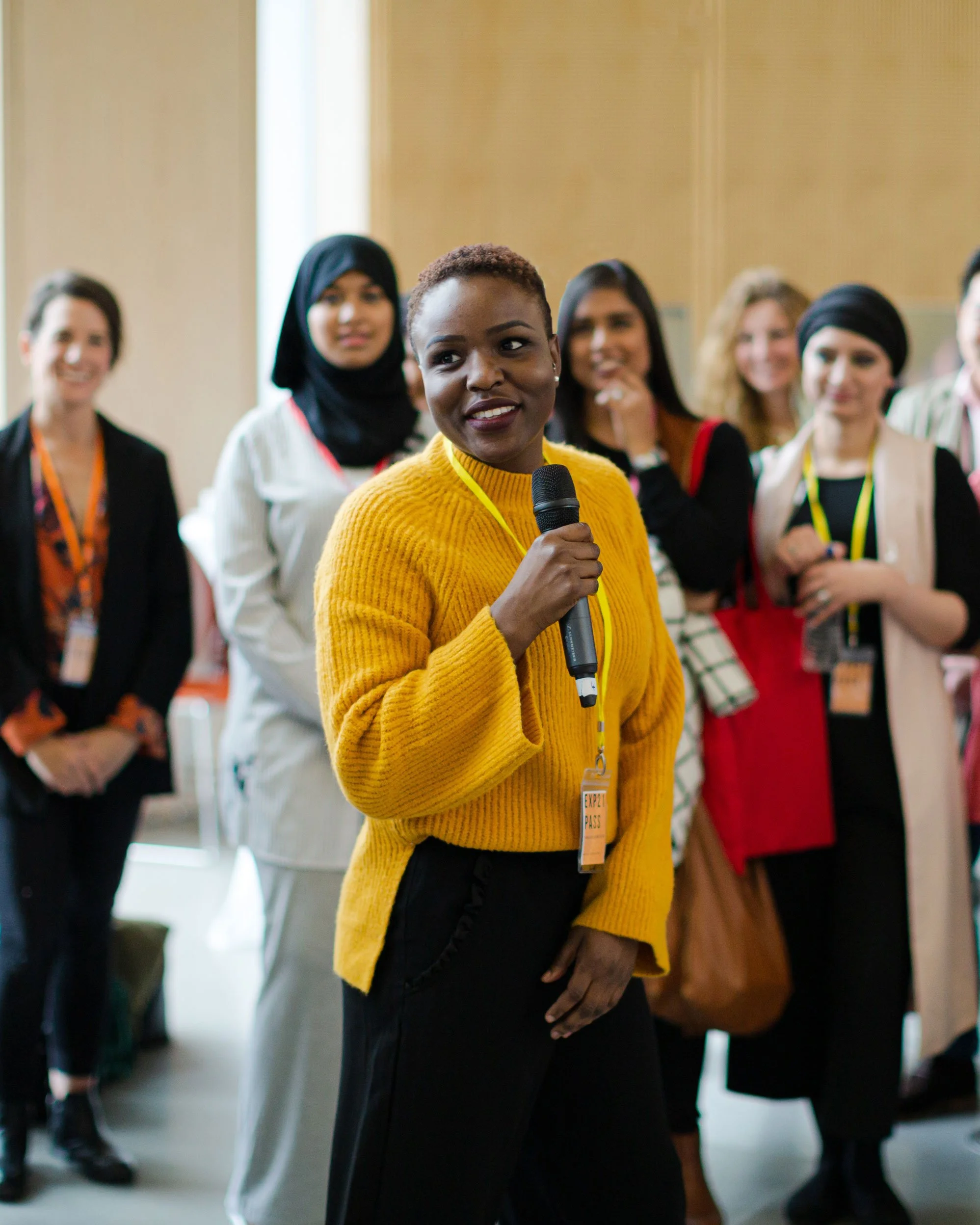 A woman in a yellow sweater holding a microphone and speaking at an indoor event with a diverse group of people listening.