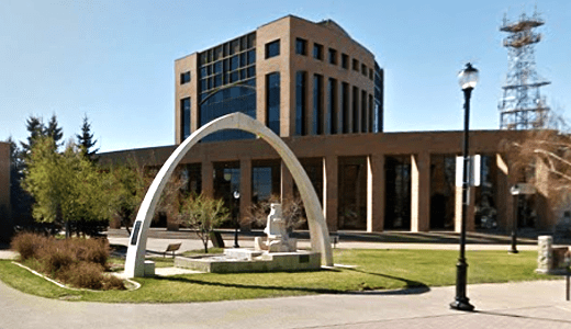 A modern building with large windows, a white arch structure in front, and a grassy area with trees and benches.